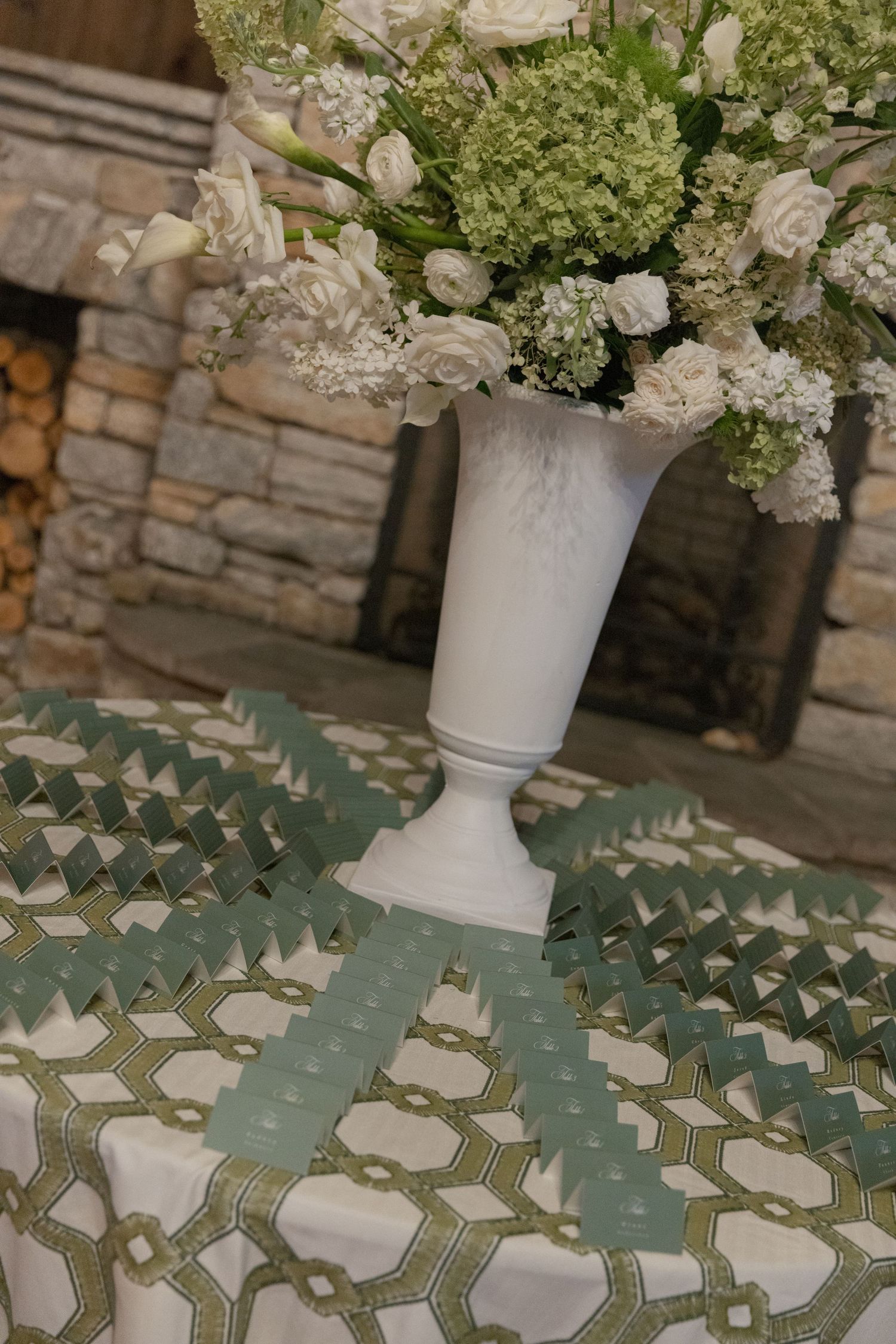 White hydrangeas and roses in a tall white urn sit above green escort cards arranged on a patterned tablecloth by a stone fireplace.