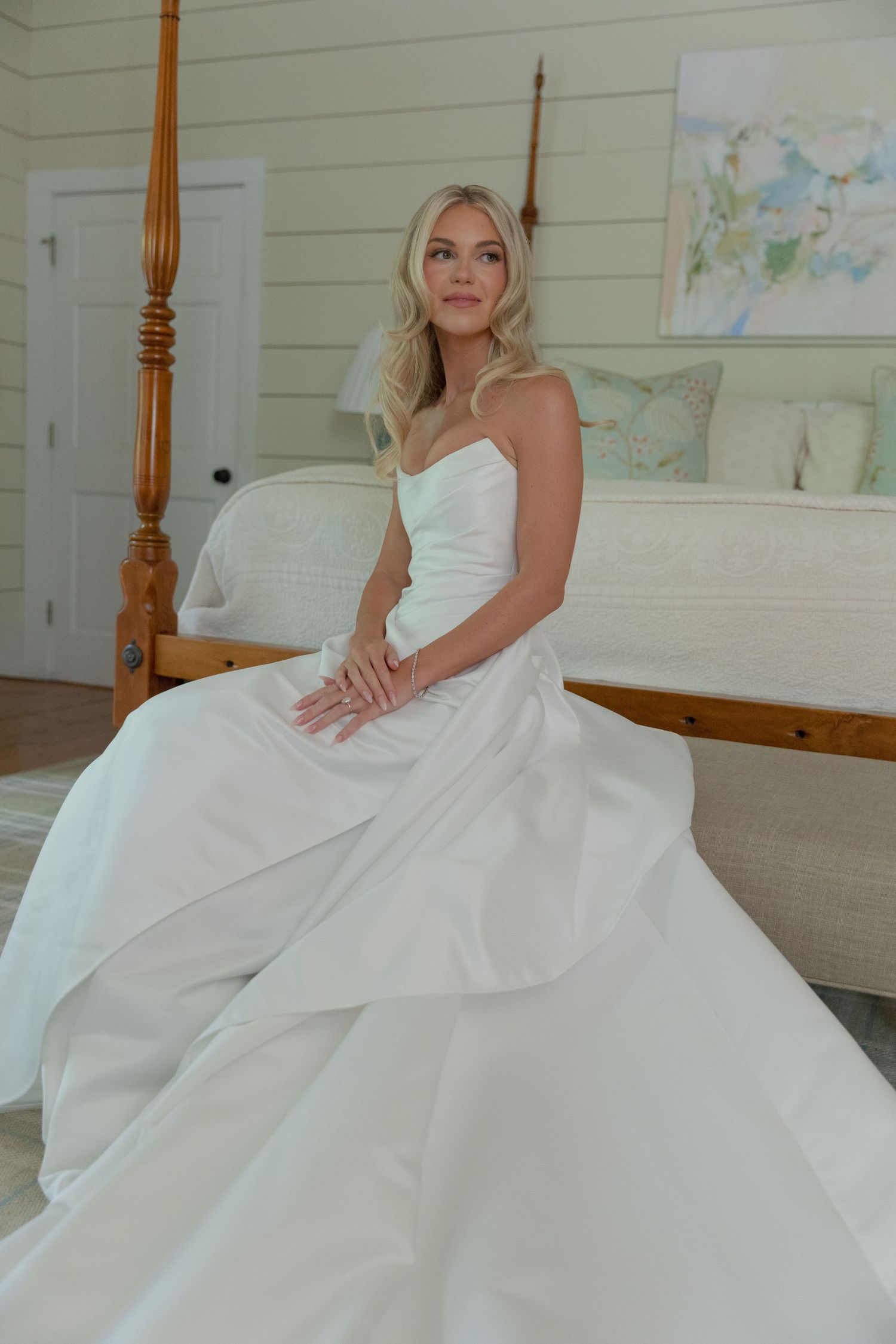 Smiling bride in a strapless white satin ballgown sits beside a wooden four-poster bed in a bright, airy bedroom.