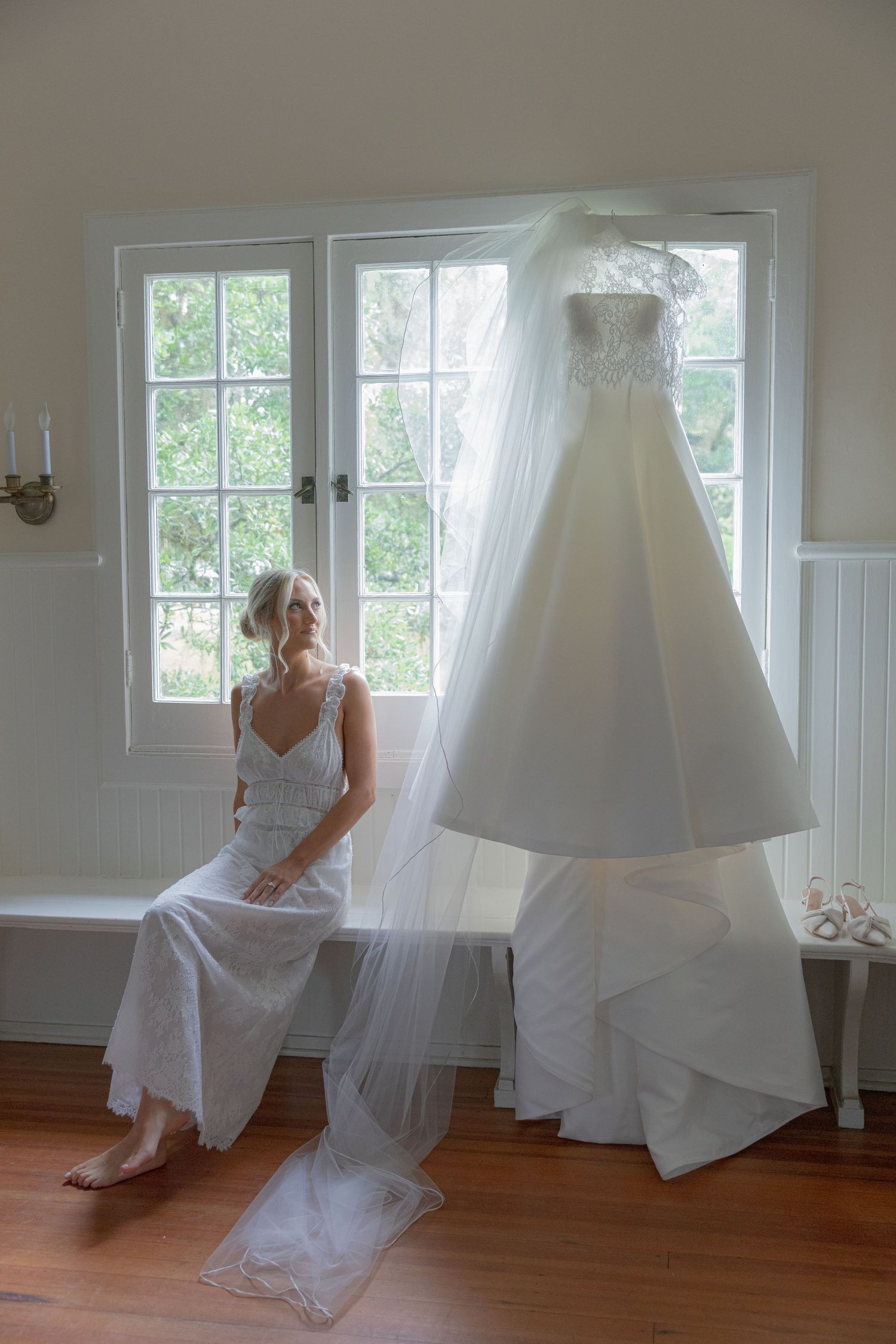 Bride in slip dress sits gazing at her lace wedding gown hanging by sunlit window in elegant bridal suite.