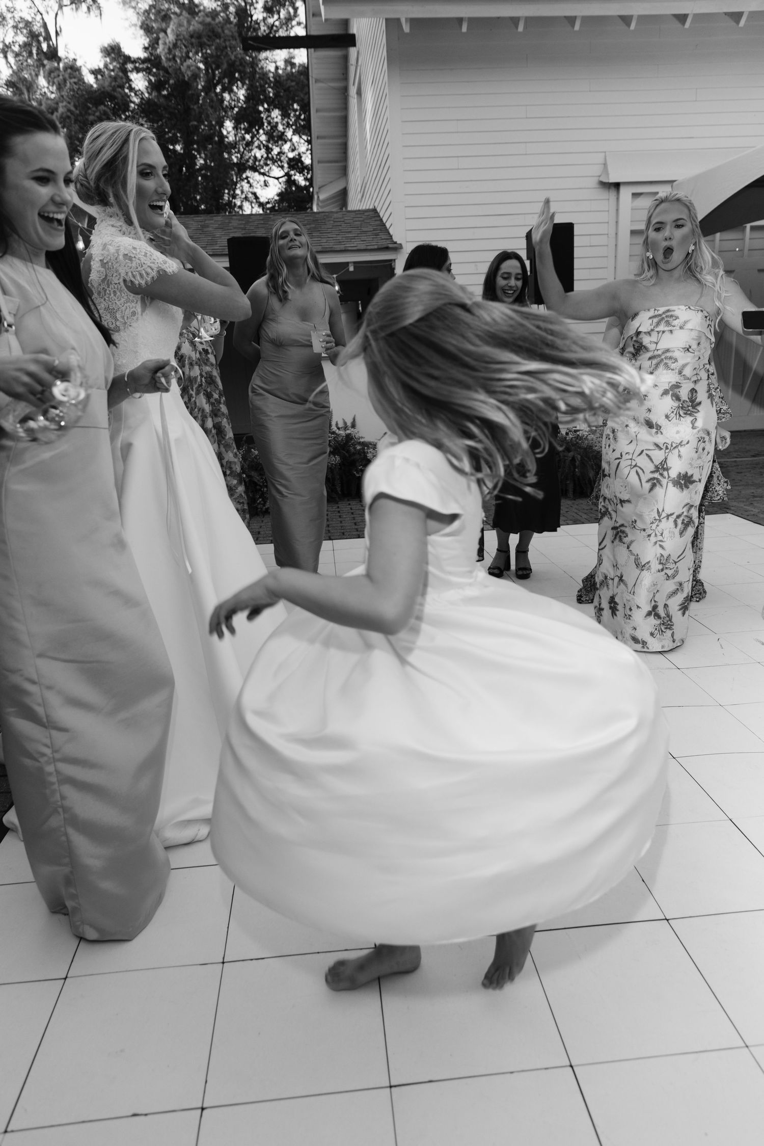 Black and white photo of flower girl twirling on dance floor surrounded by laughing bridesmaids at outdoor reception.