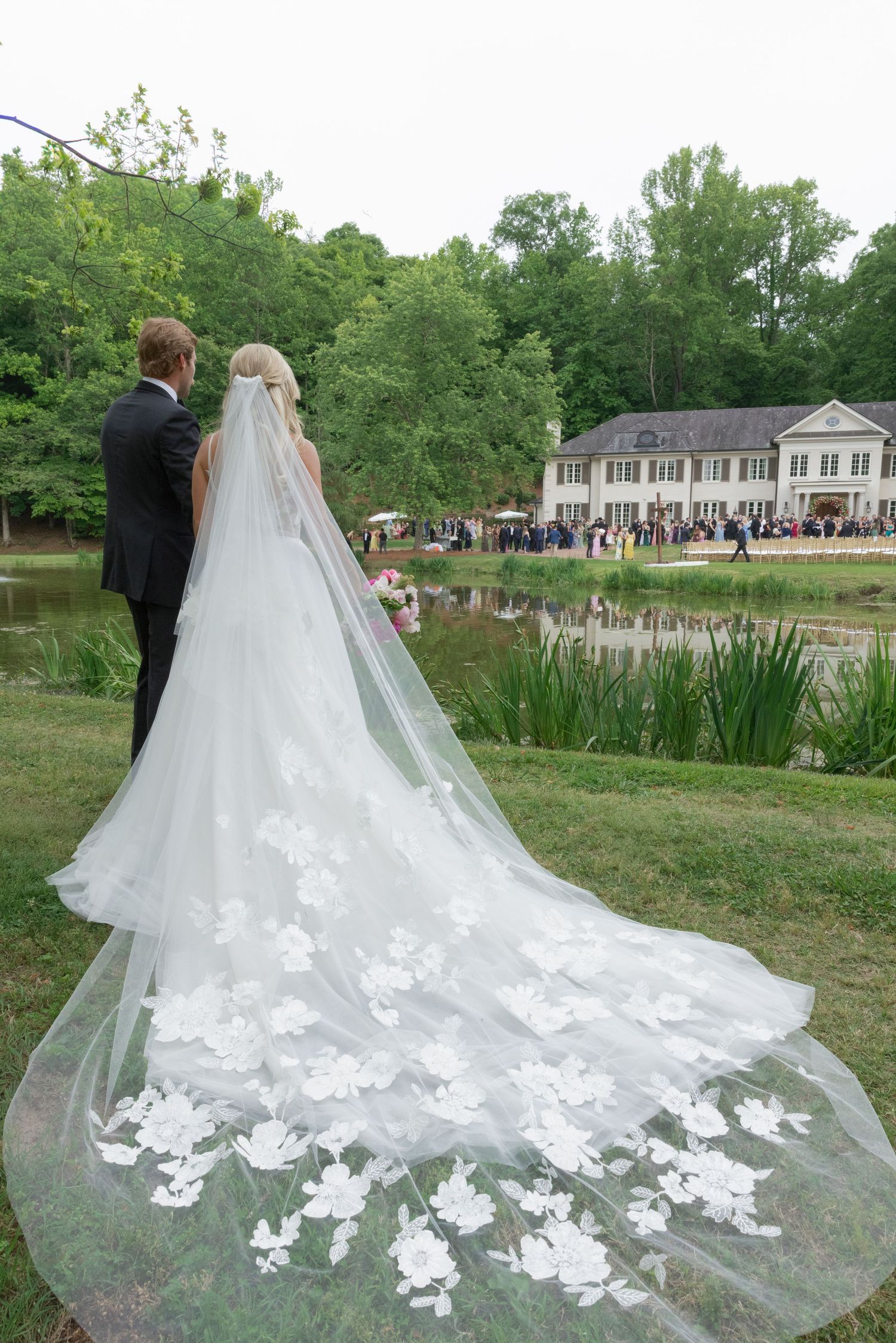 Bride and groom stand overlooking a pond with guests gathered at an elegant venue, bride wearing a floral lace cathedral veil.
