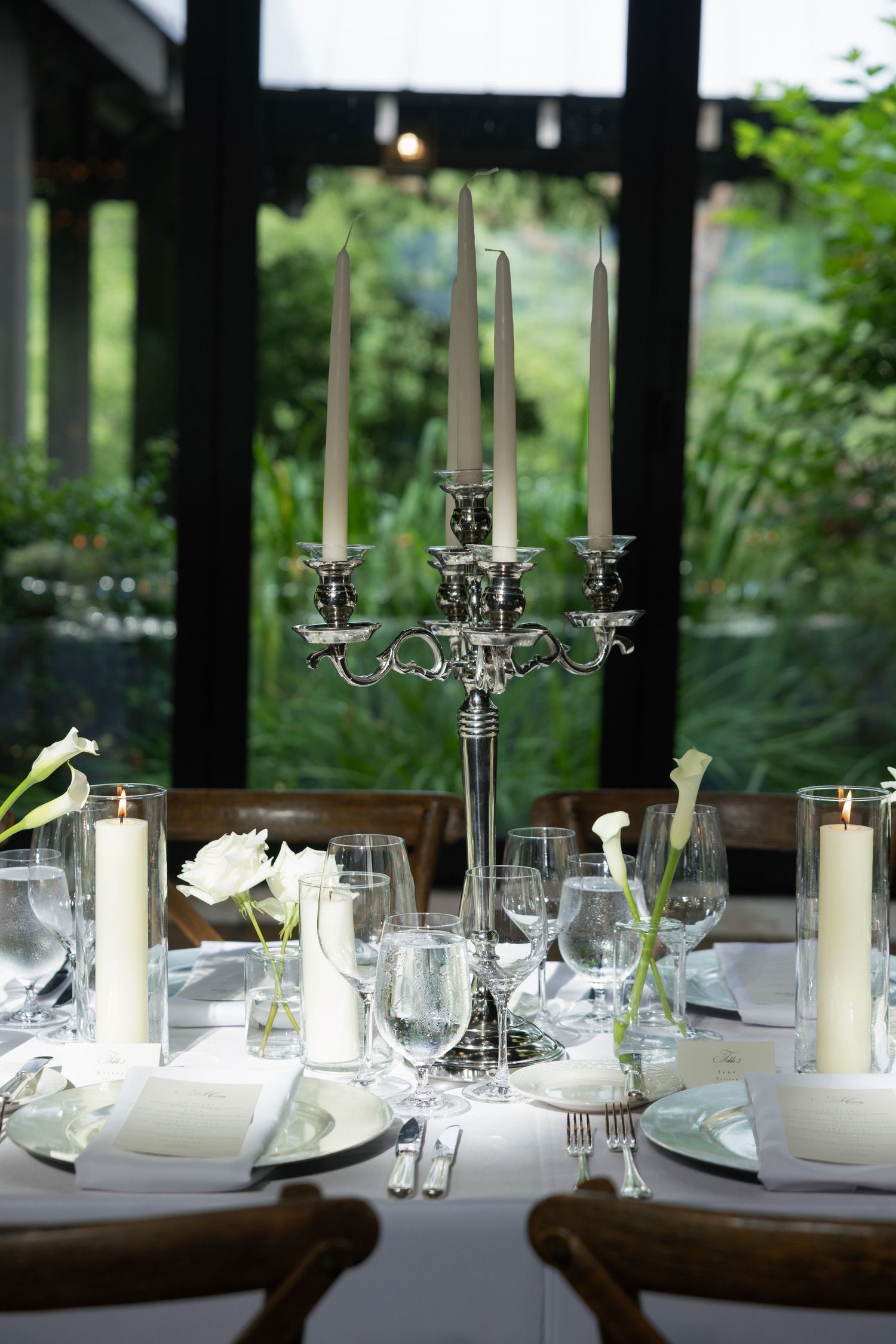 Elegant wedding table setting with a silver candelabra, white flowers, pillar candles and crystal glassware beside large garden windows.