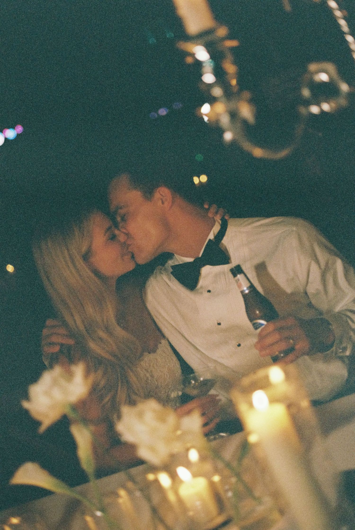 A bride and groom in a bow tie share a kiss at a candlelit reception table with white flowers and a chandelier overhead.