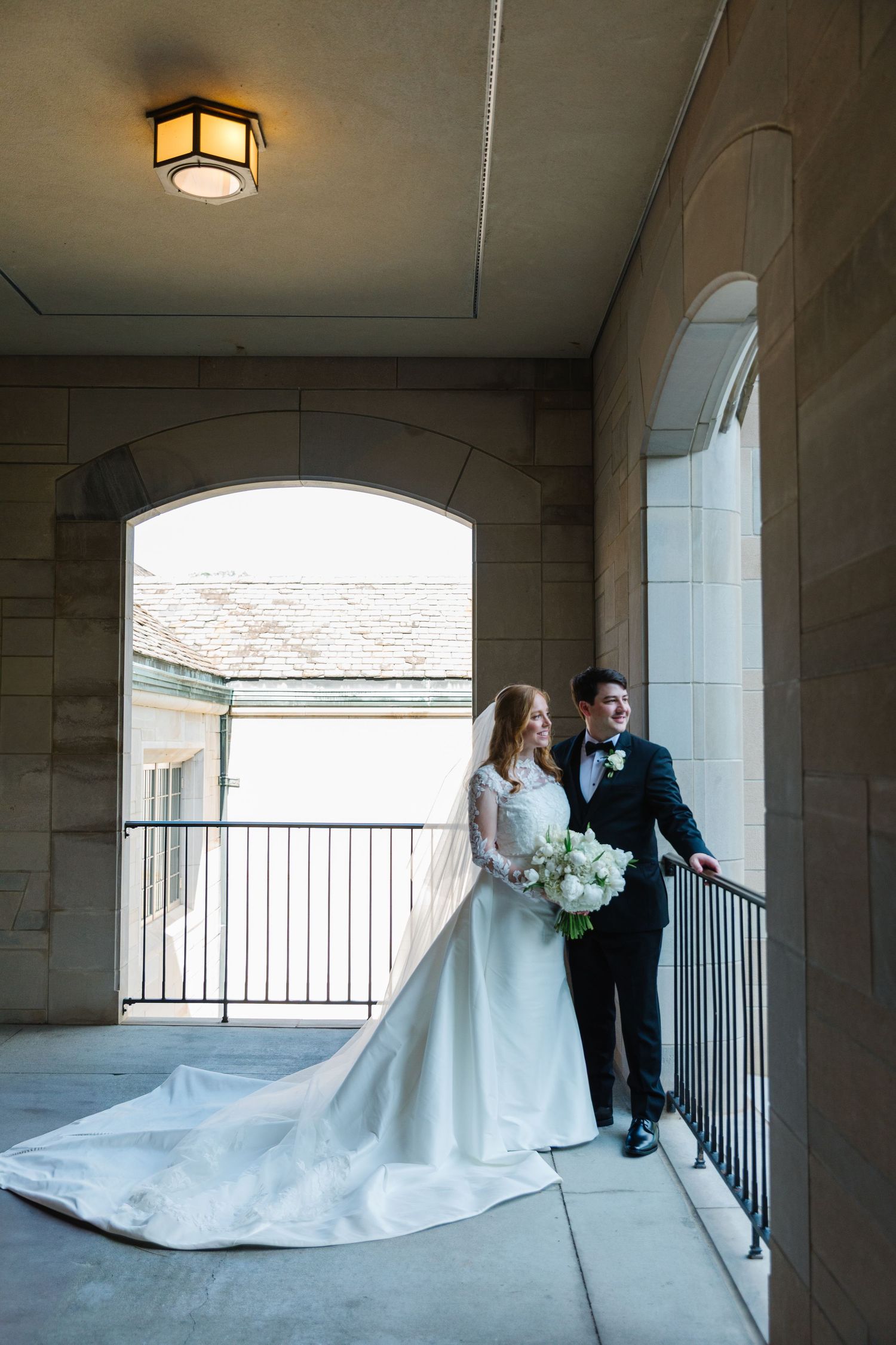A wedding couple poses together in an elegant archway corridor with natural light streaming in from windows.