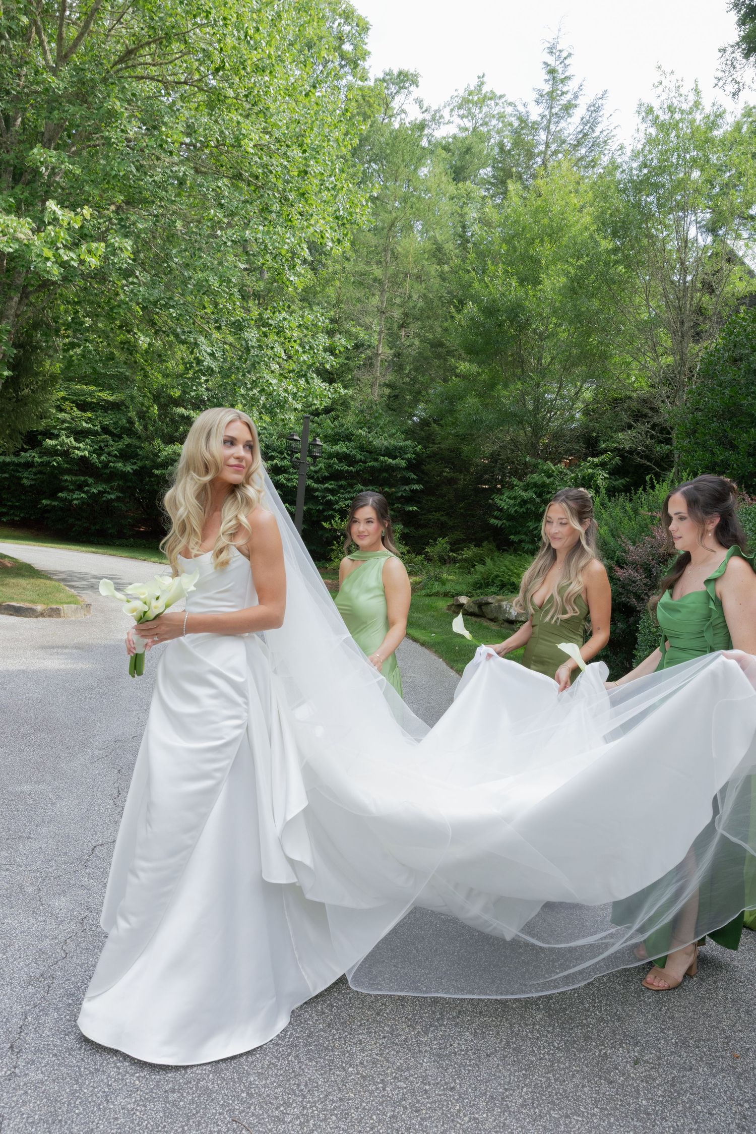 Bride in a white satin gown and long veil holds a bouquet while bridesmaids in green dresses hold her train outdoors among trees.