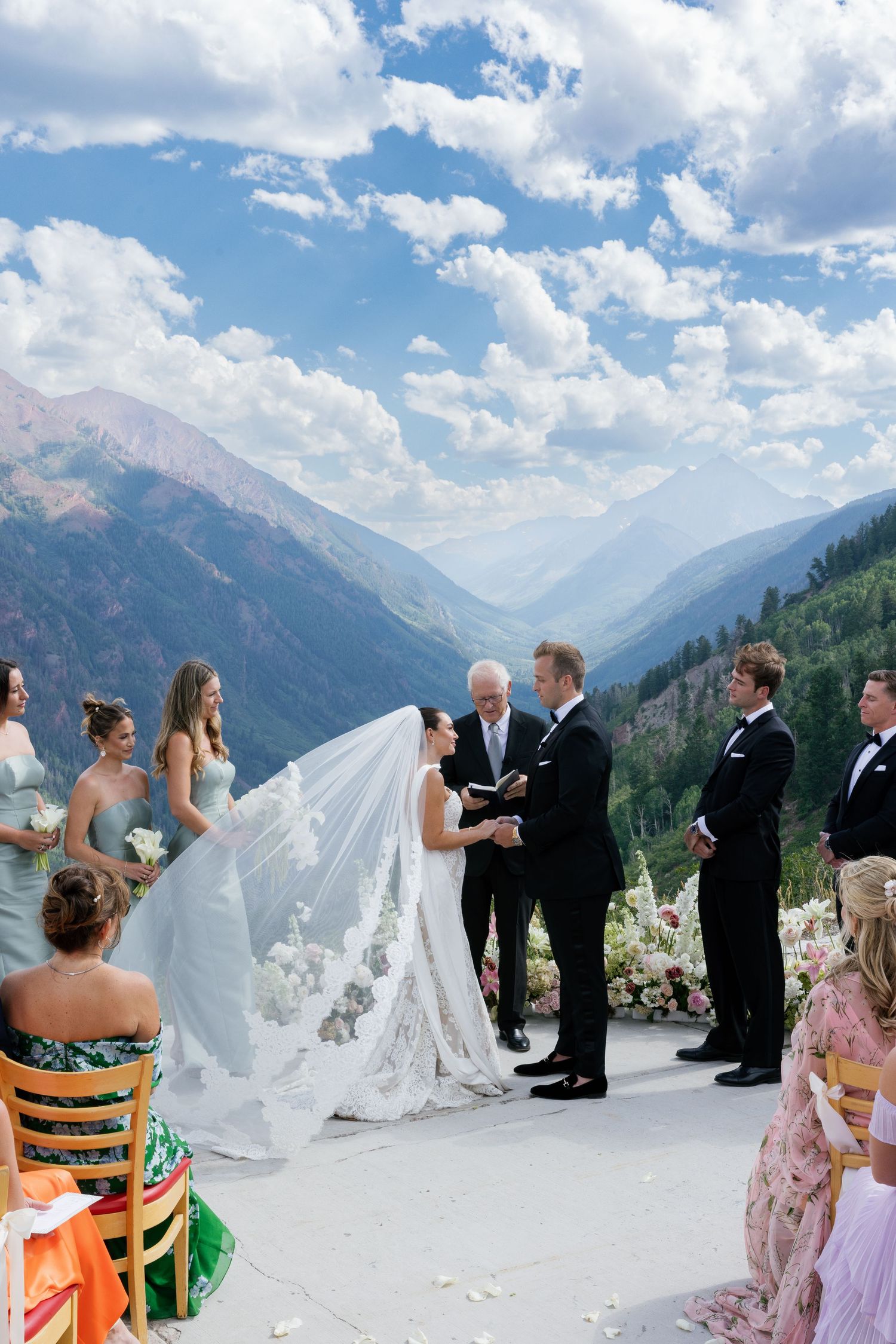 A scenic mountain wedding ceremony takes place on a high-altitude deck with dramatic snow-capped peaks in the background.