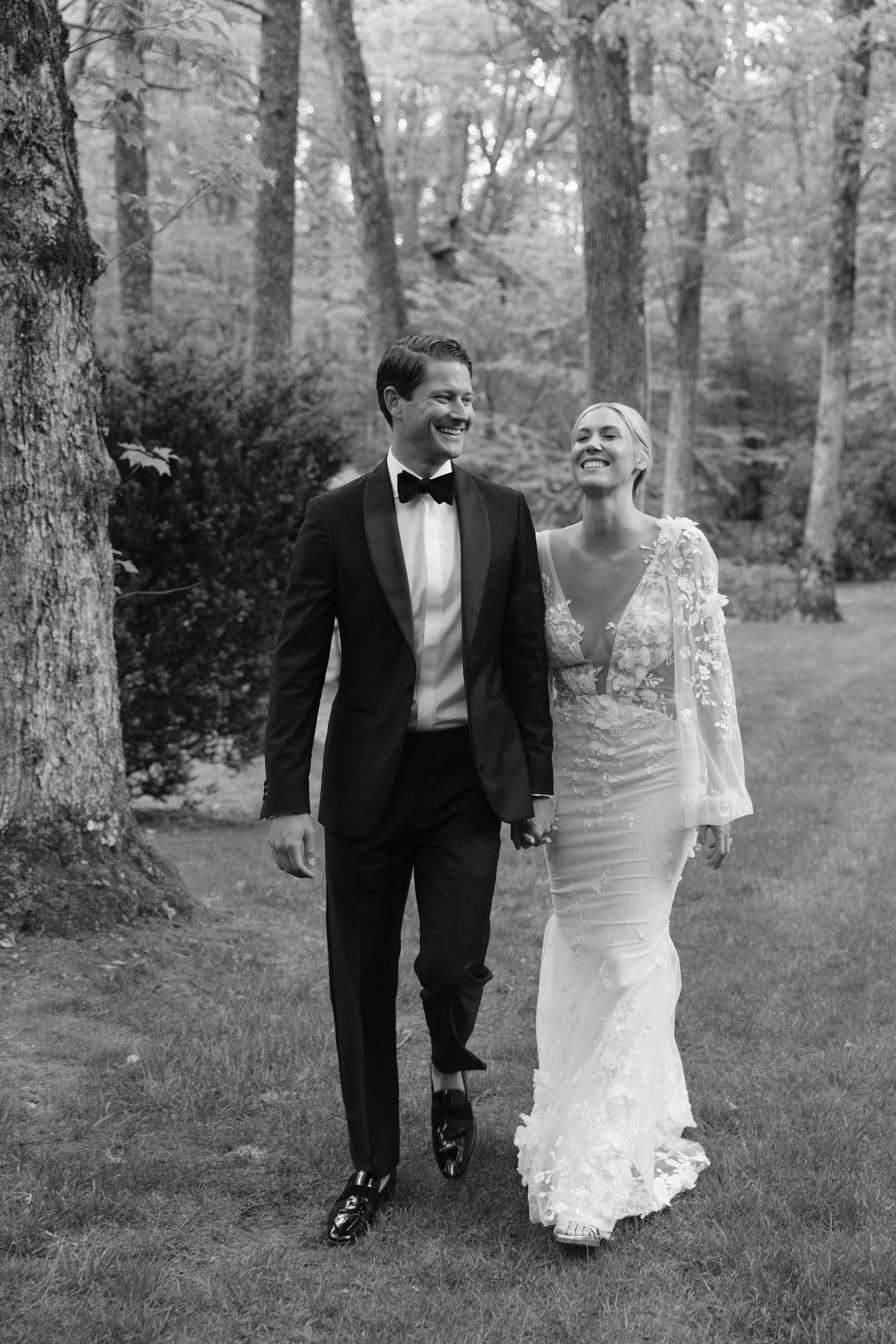 A black and white photo of a couple walking and laughing together in a wooded outdoor setting.
