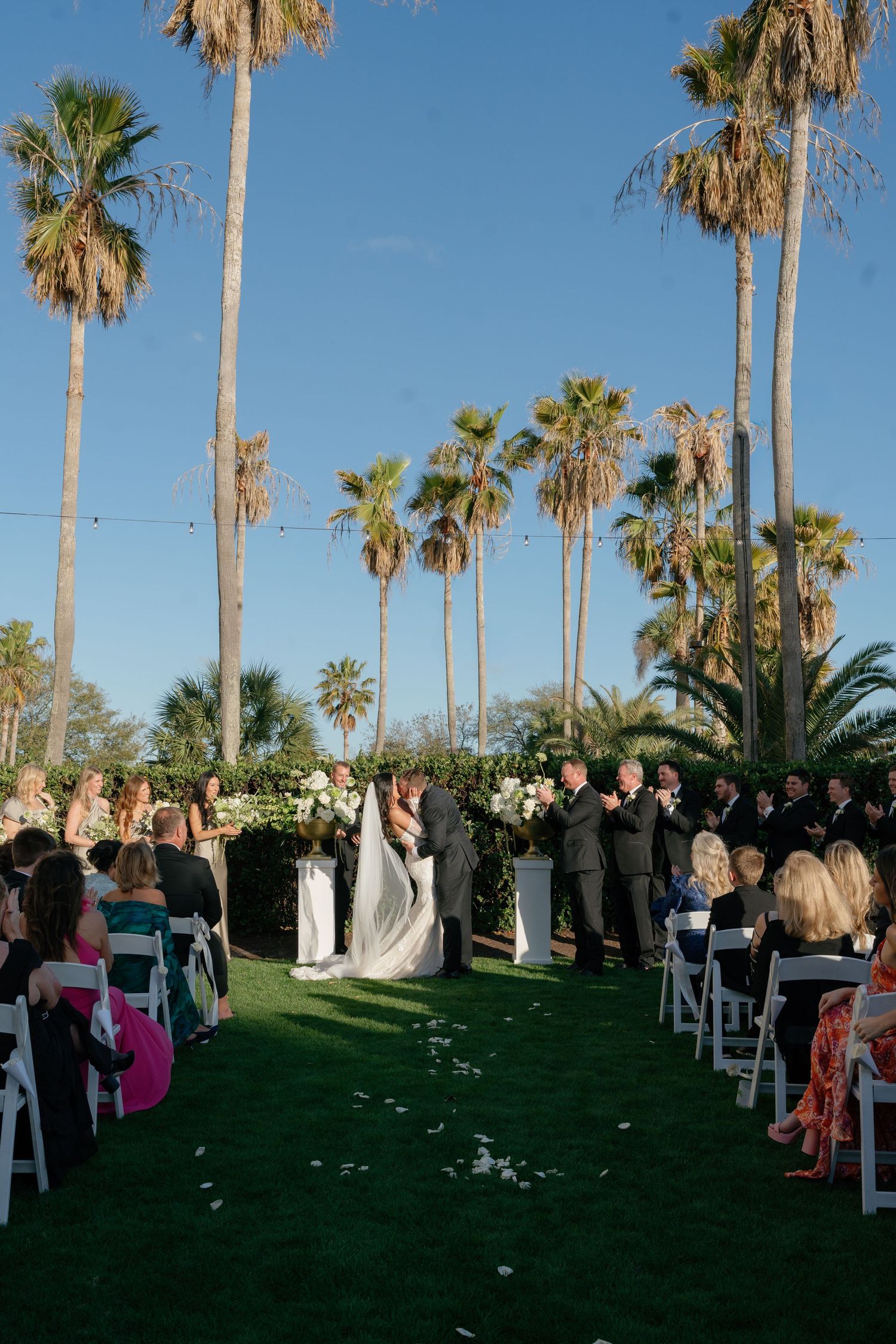 Outdoor wedding ceremony on green lawn with tall palm trees, white chairs, and couple at floral altar under clear blue sky.