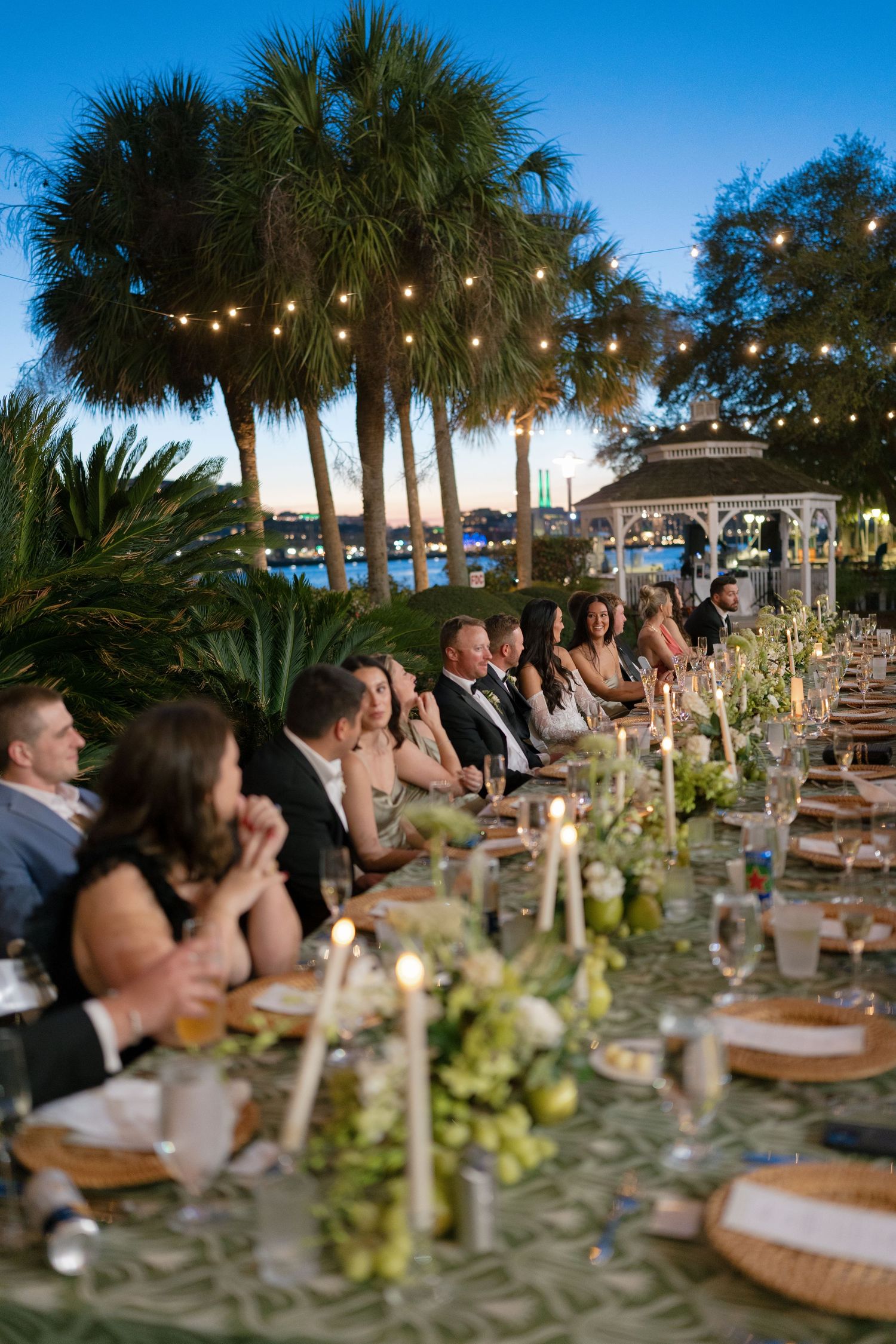 Elegant outdoor wedding reception at dusk with string lights, palm trees, candles, and guests seated at a long decorated table.
