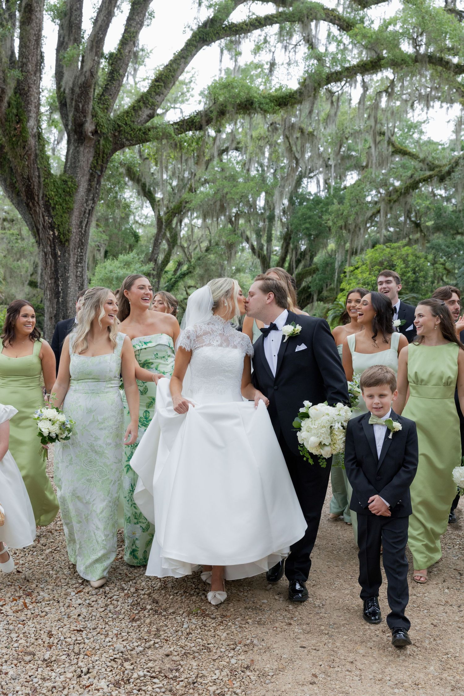 Bride and groom kissing while walking with wedding party in sage green dresses under Spanish moss-draped oak trees.