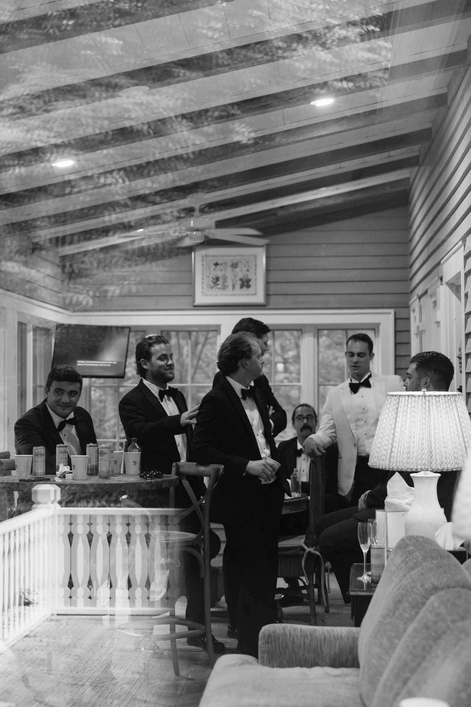 Black and white photo of groomsmen in tuxedos and bow ties chatting and sharing drinks on a cozy covered porch before the wedding.