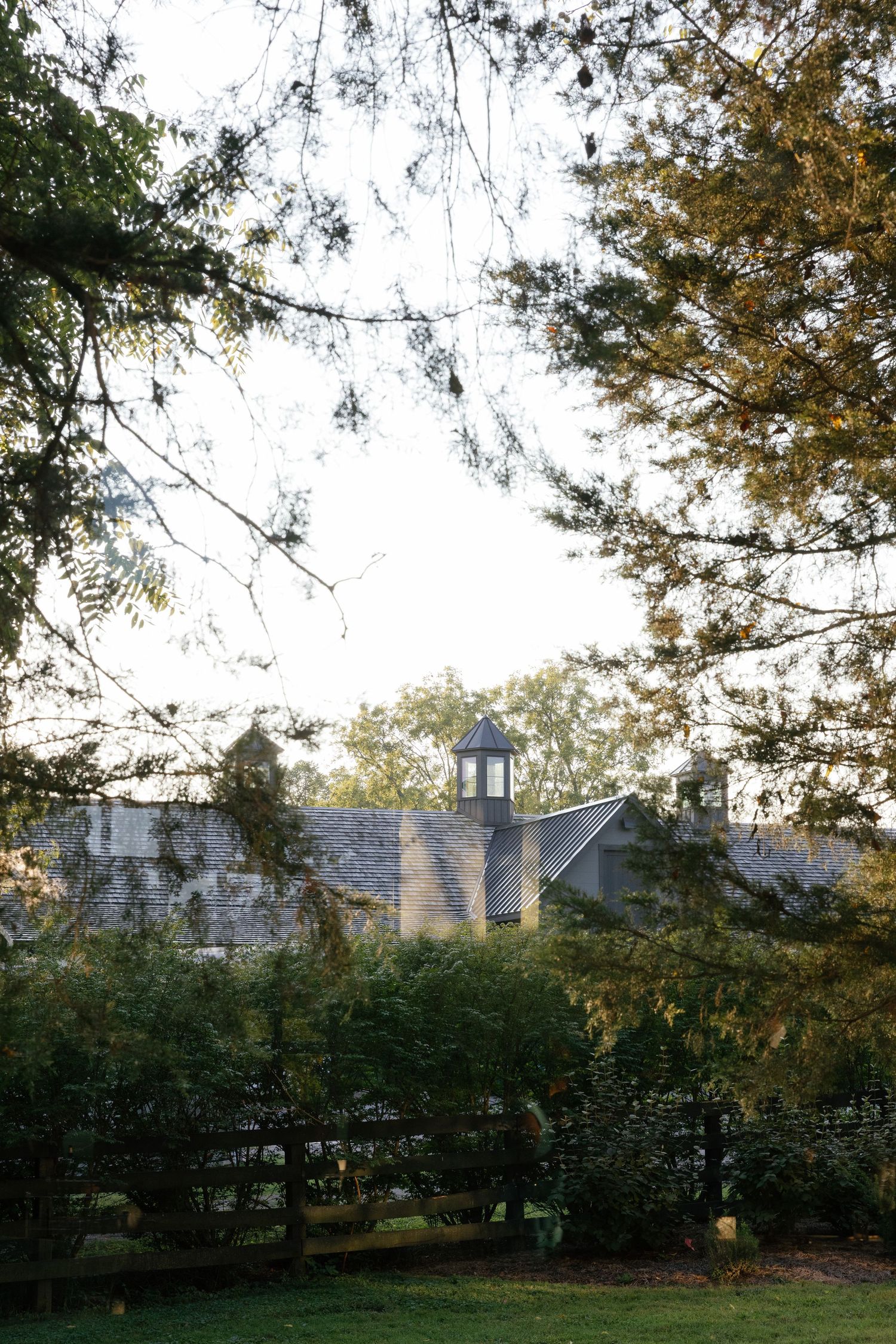 A rustic barn with a white cupola peeks through autumn trees and foliage against an overcast sky.