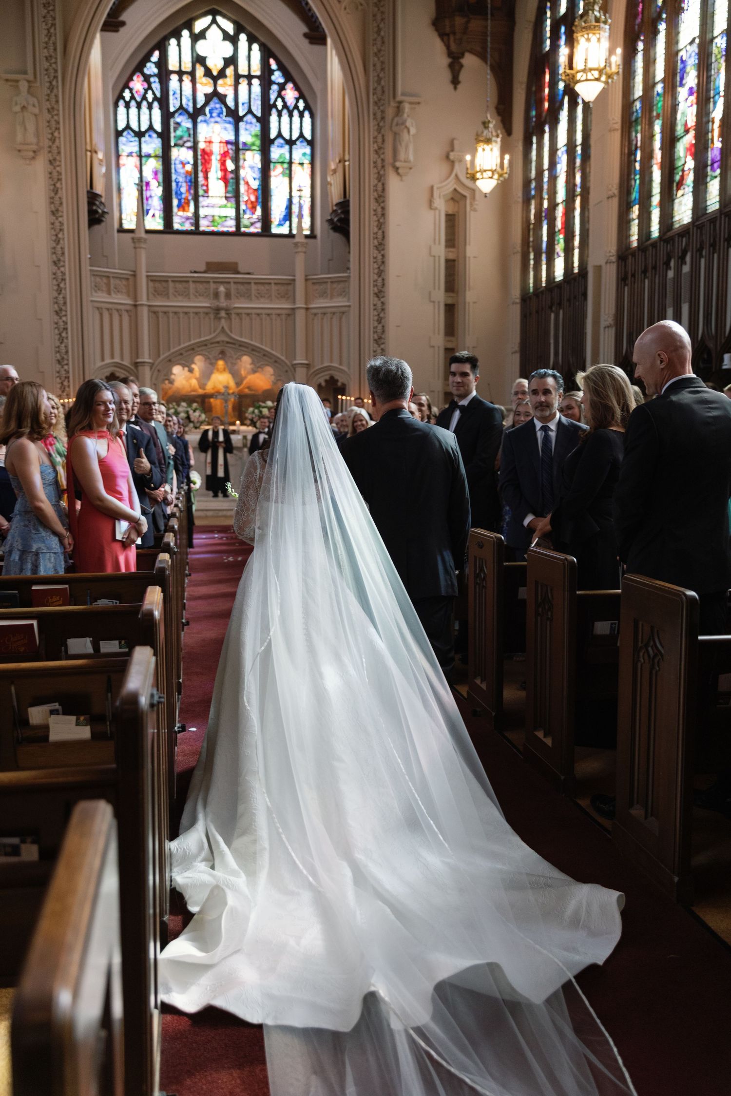 A bride in a flowing white wedding gown with a long train walks down the aisle of a gothic church with stained glass windows.