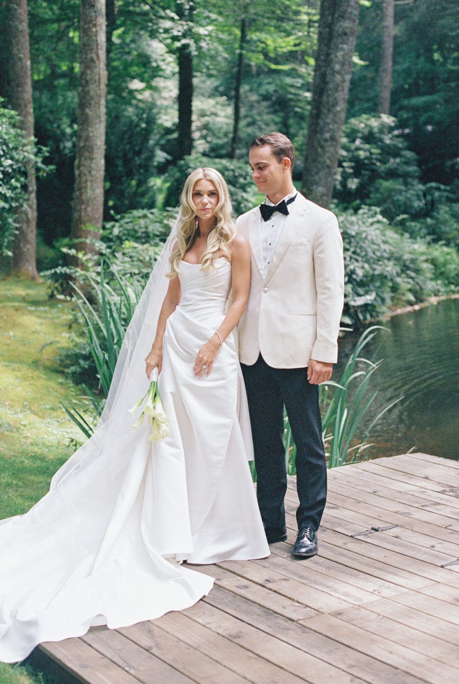 Bride in a strapless white gown with a long veil and groom in a white dinner jacket and bow tie pose on a wooden dock by a pond.