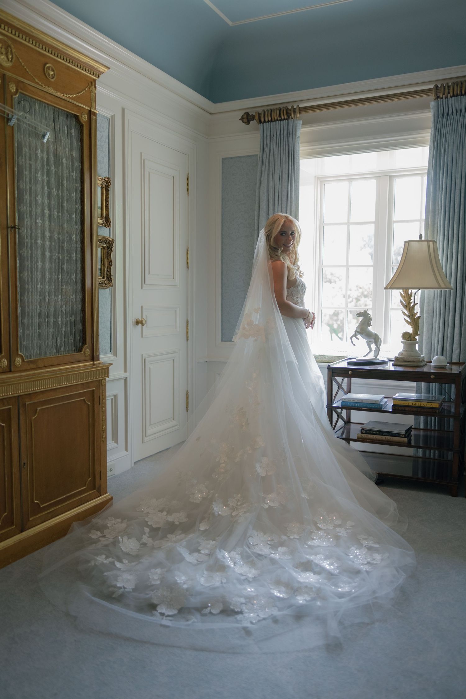 Bride in a stunning gown with floral lace cathedral veil stands by a sunlit window in an elegant blue and gold room.