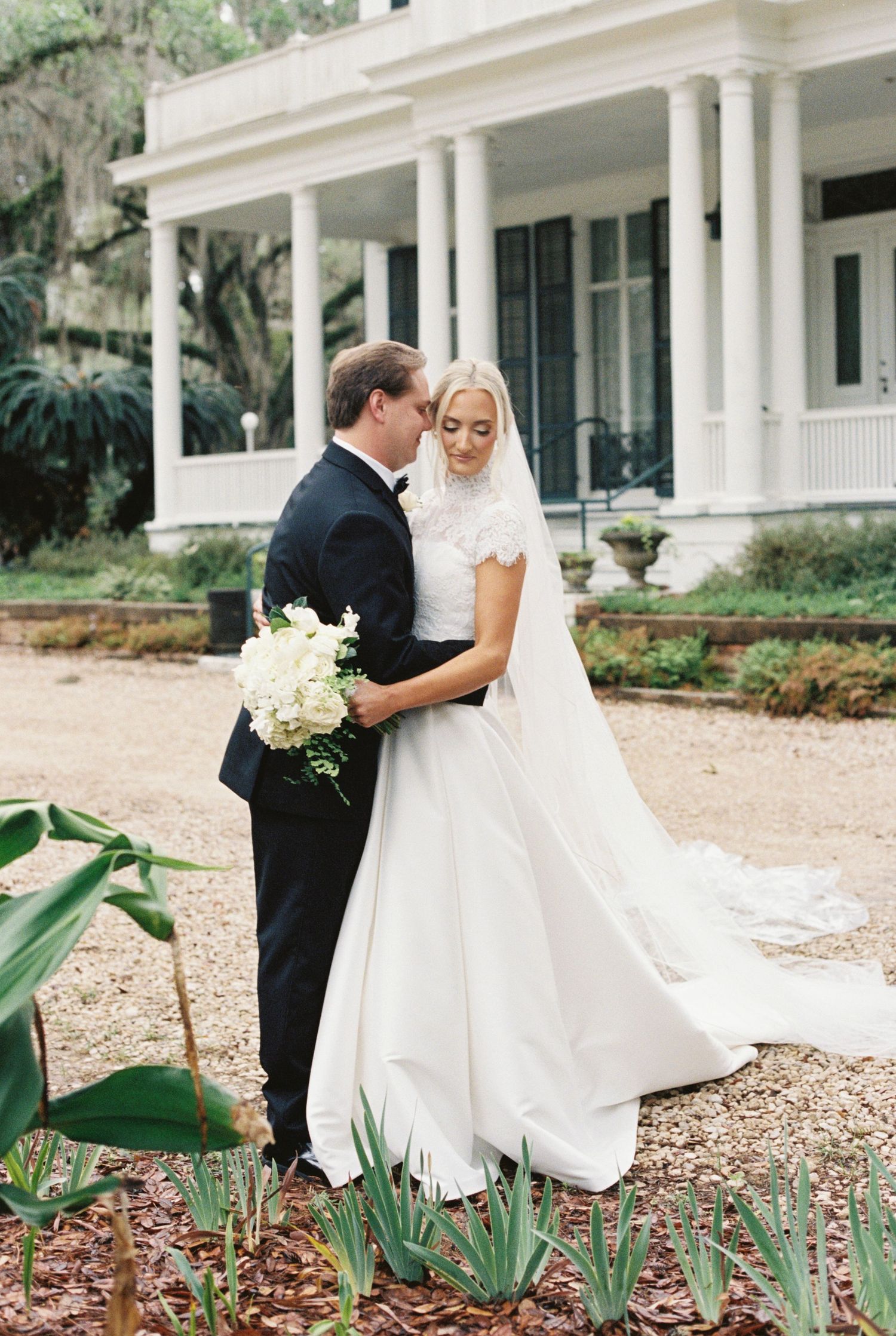 Bride in white gown with veil and groom in black tuxedo embrace outside a classic Southern plantation home.