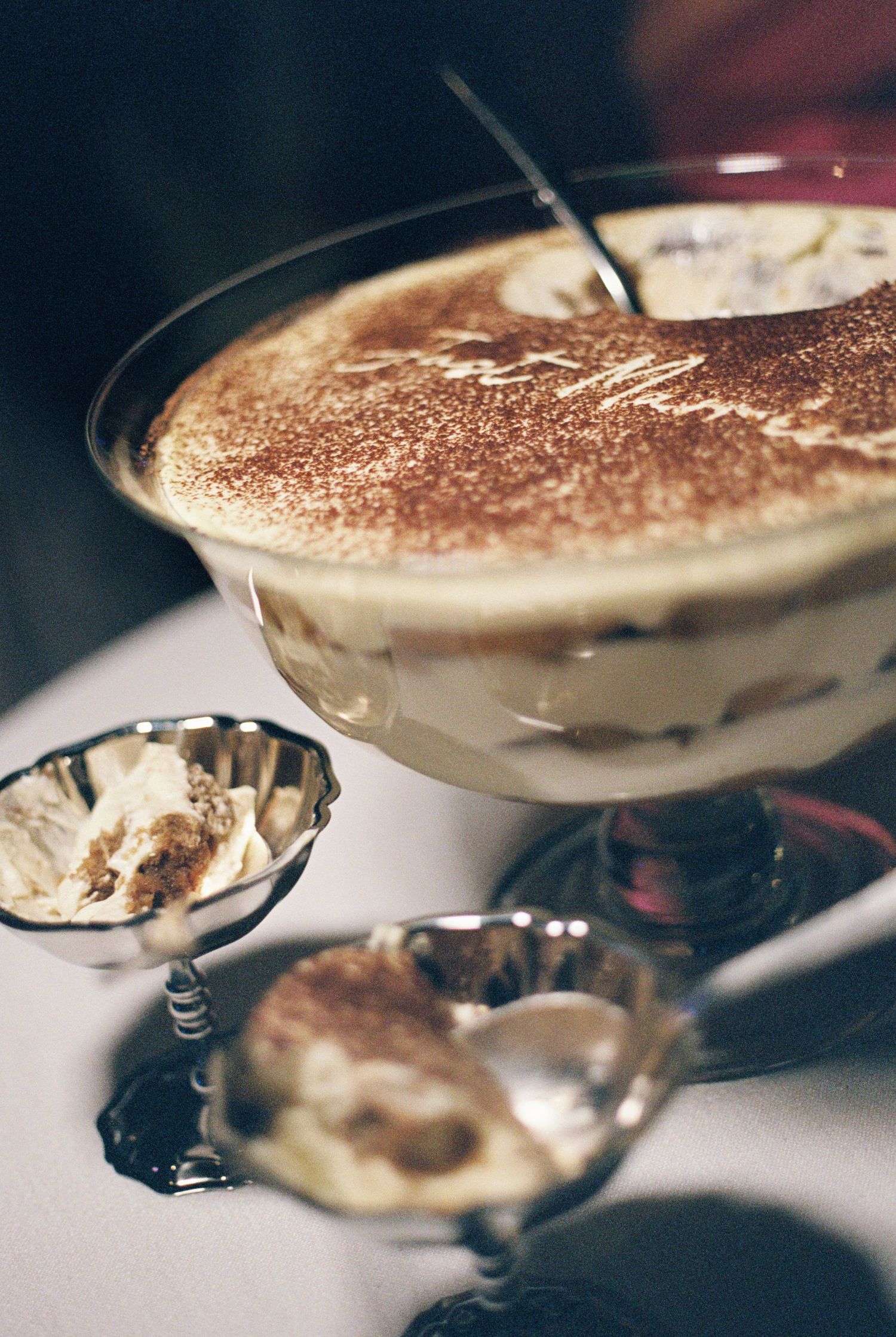 Layered tiramisu dusted with cocoa powder in a large pedestal glass bowl, served with two small silver dessert dishes on a table.