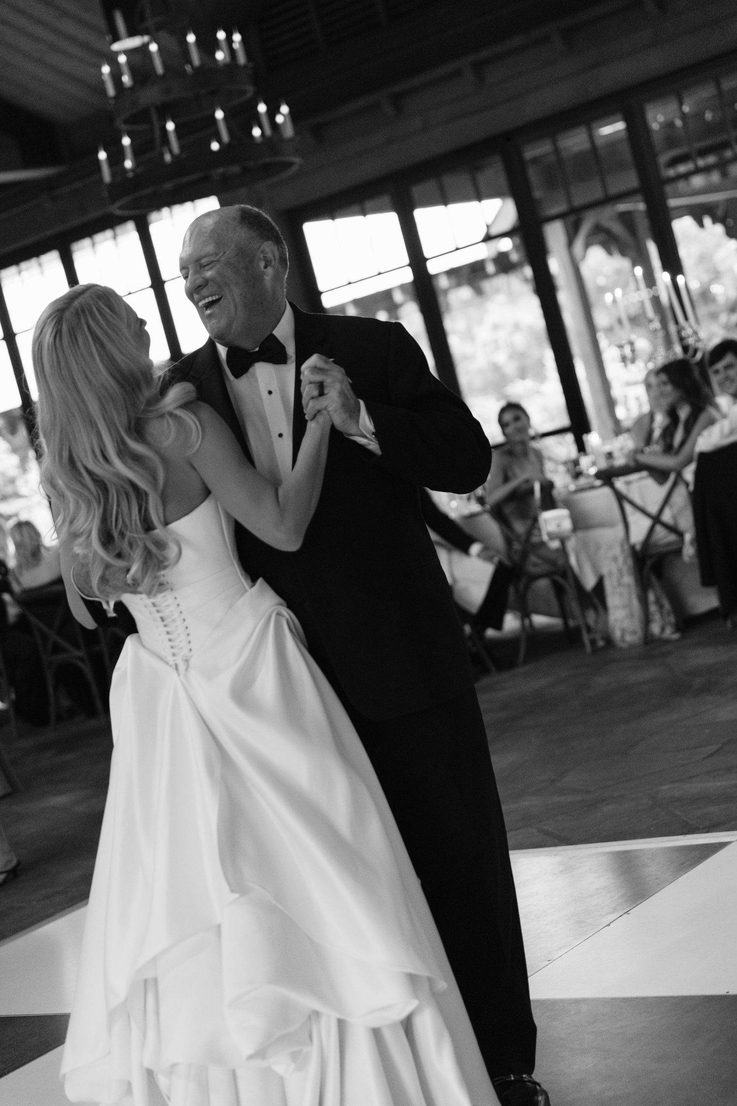 Black and white photo of a bride in a lace-up corset gown dancing and laughing with a man in a tuxedo as guests look on.