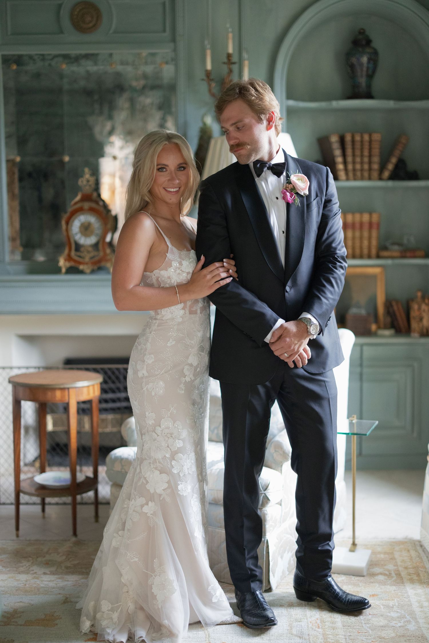 Elegant bride in lace gown stands beside groom in black tuxedo with pink boutonniere in a classic interior.