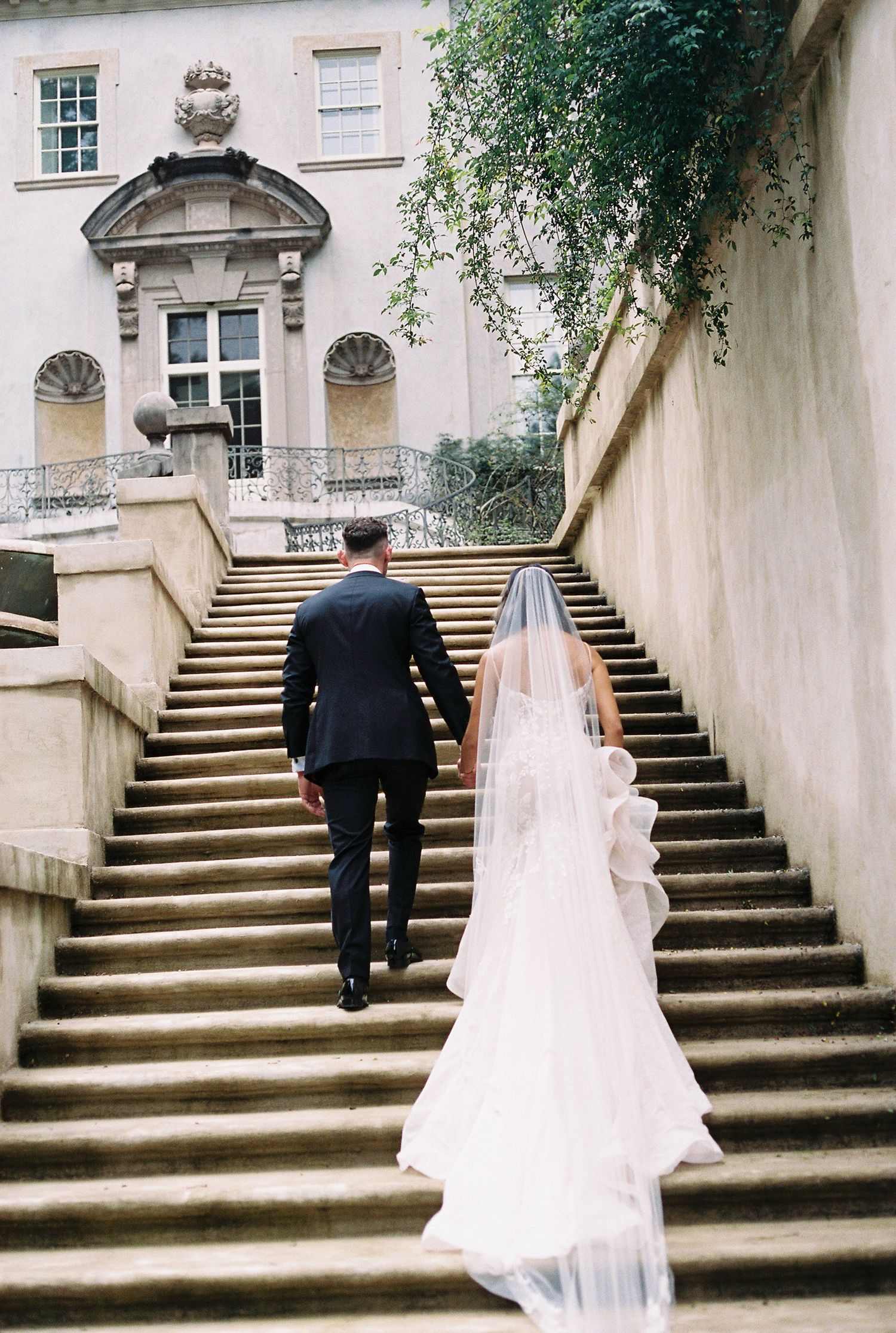 A couple in formal wedding attire ascending grand stone steps of an elegant historic building with classical architecture. swann house