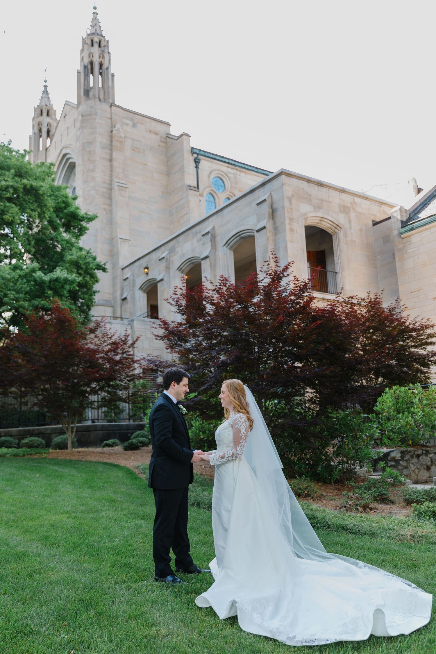 Wedding couple stands together on church grounds with Gothic architecture in background.
