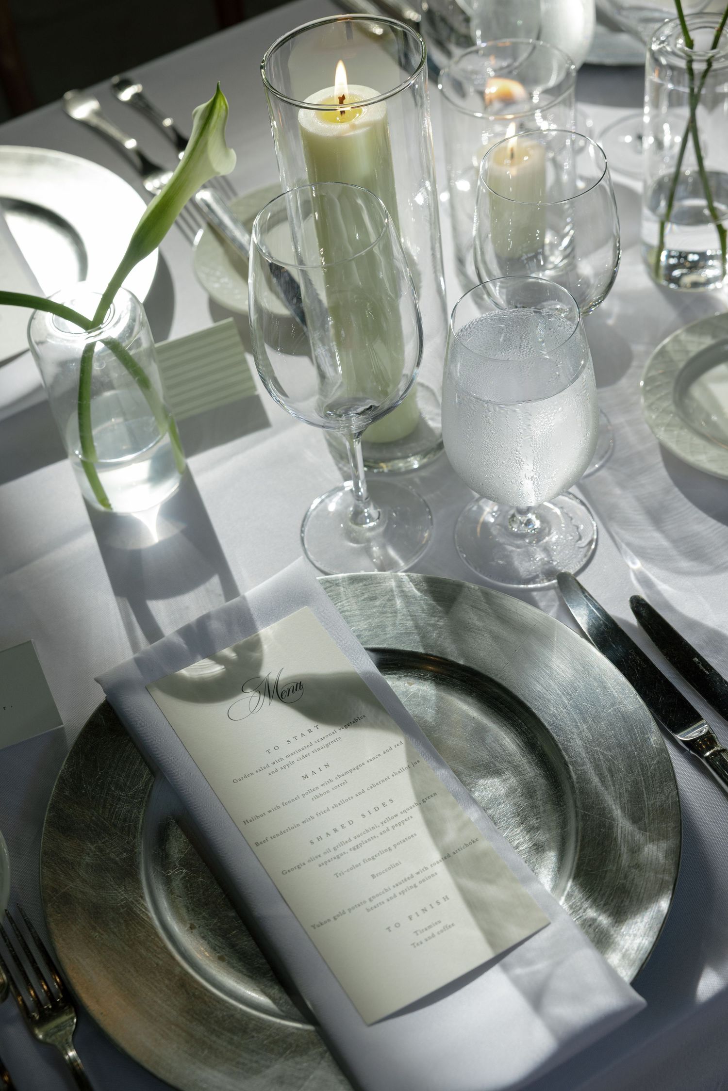 An elegant wedding table setting with a silver charger plate, folded menu, candles in glass holders, and a white flower stem.
