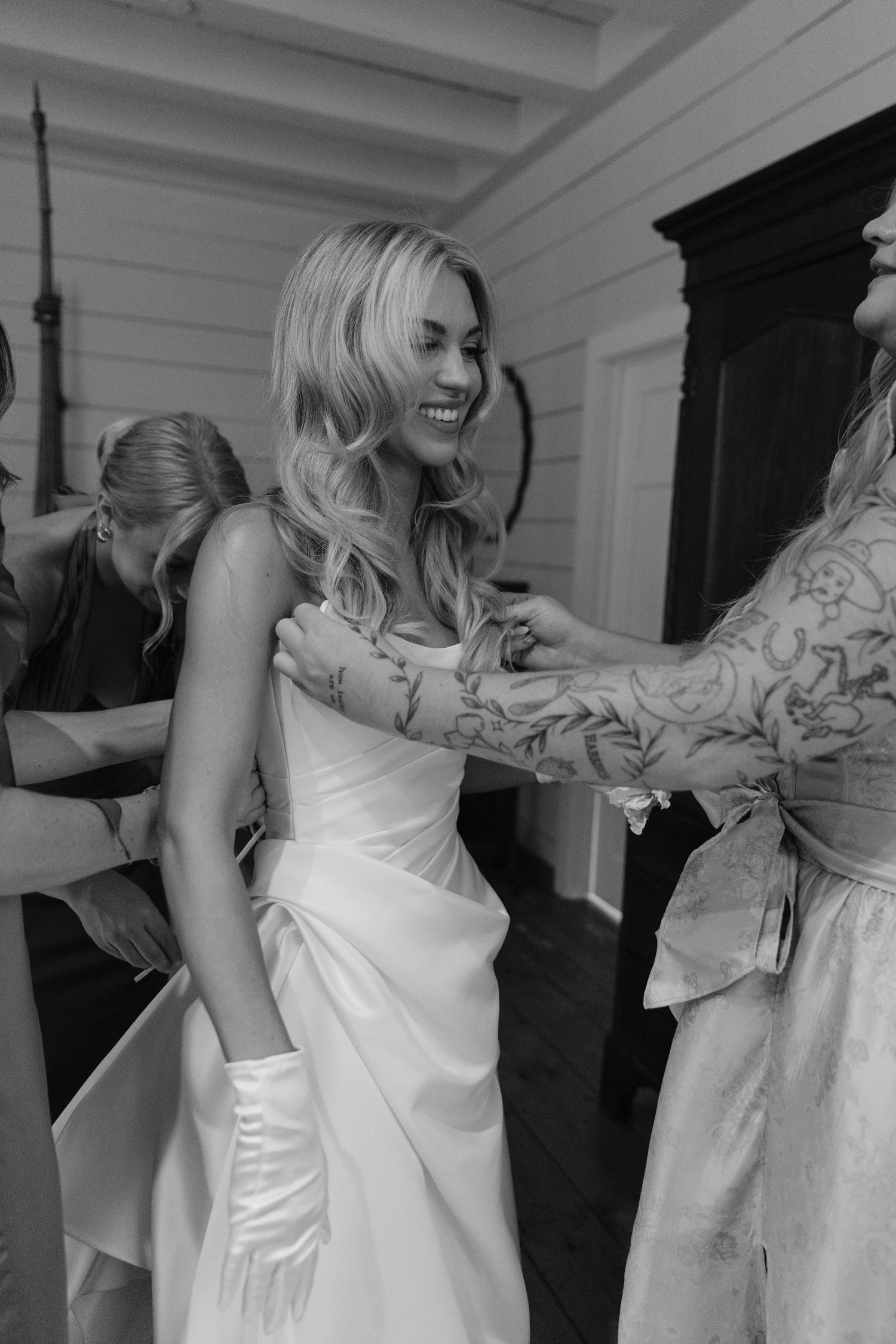 Smiling bride in a strapless satin gown and white gloves gets help with her dress from tattooed bridesmaids in a bright room.