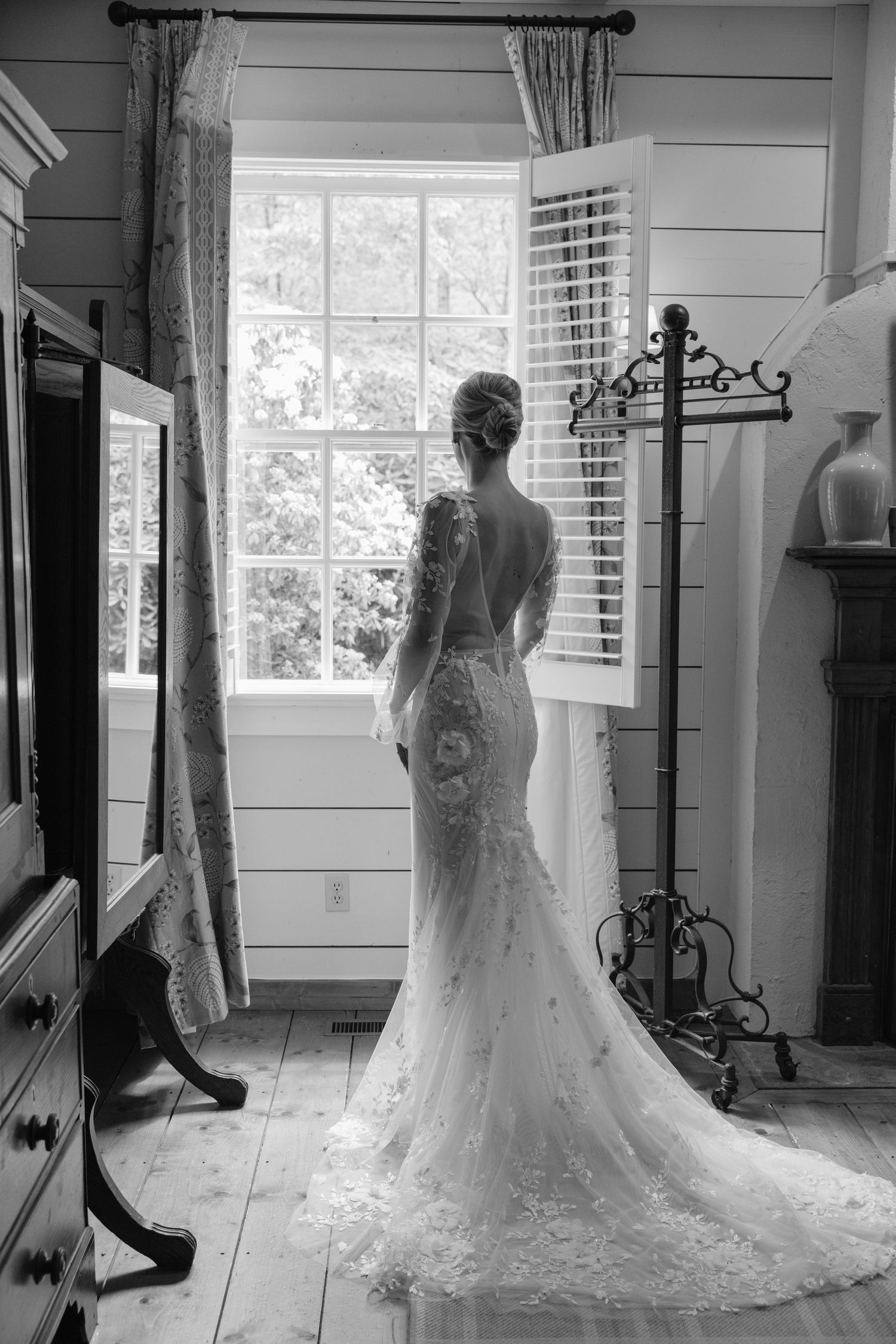 A bride in a fitted lace wedding gown with long train stands silhouetted at a window in black and white photography.