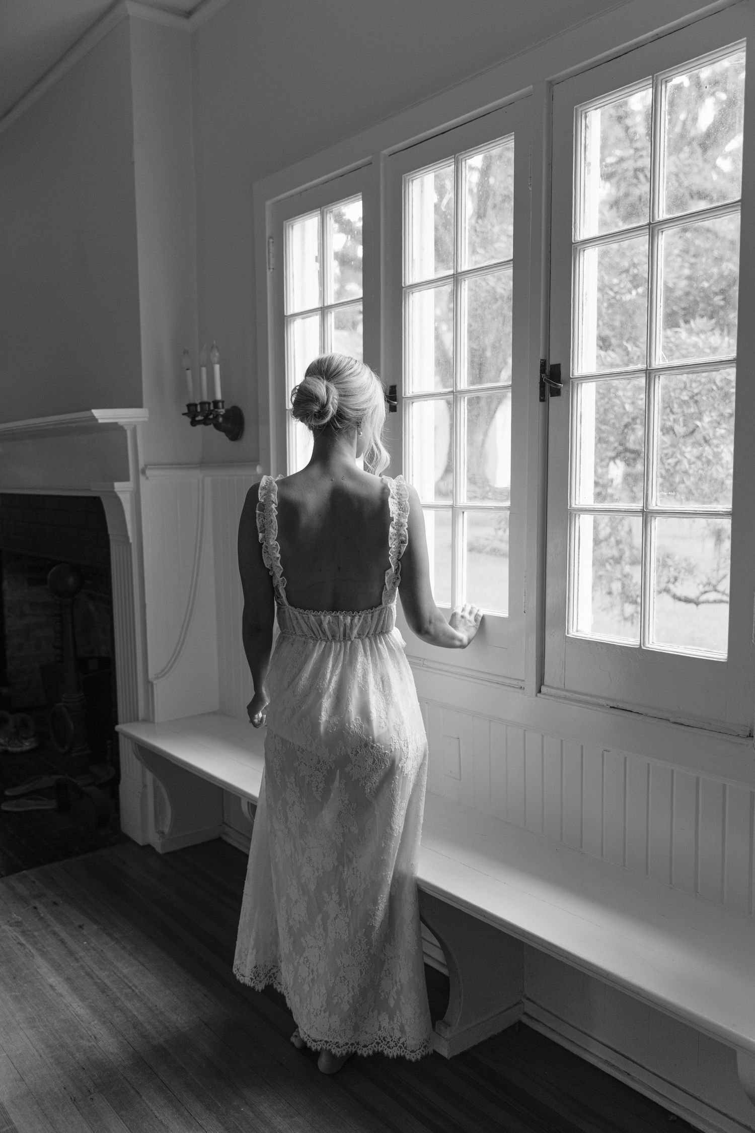 Black and white photo of a bride in a backless lace gown gazing out large windows in an elegant room.