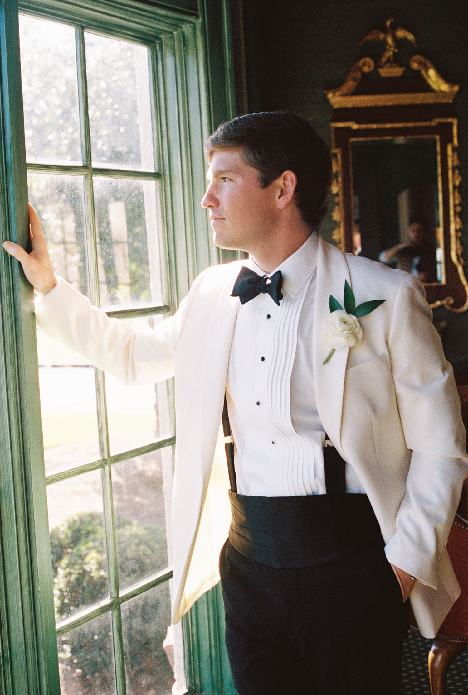A formal white tuxedo jacket and black bow tie outfit is shown against a window with green trim in elegant lighting.