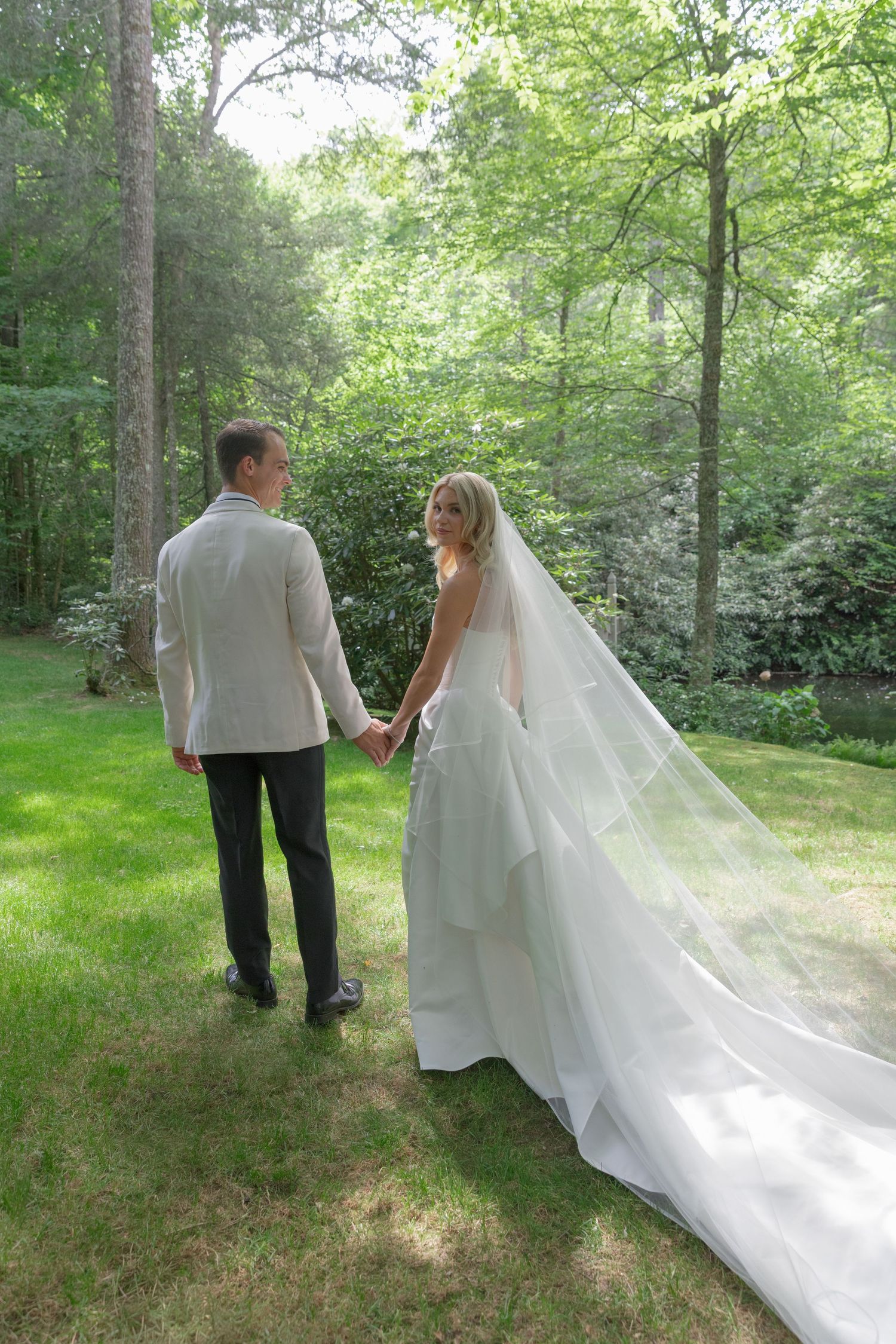 Bride in a white gown with a long veil holds hands with a groom in a light jacket while walking across a sunlit lawn in a wooded garden.