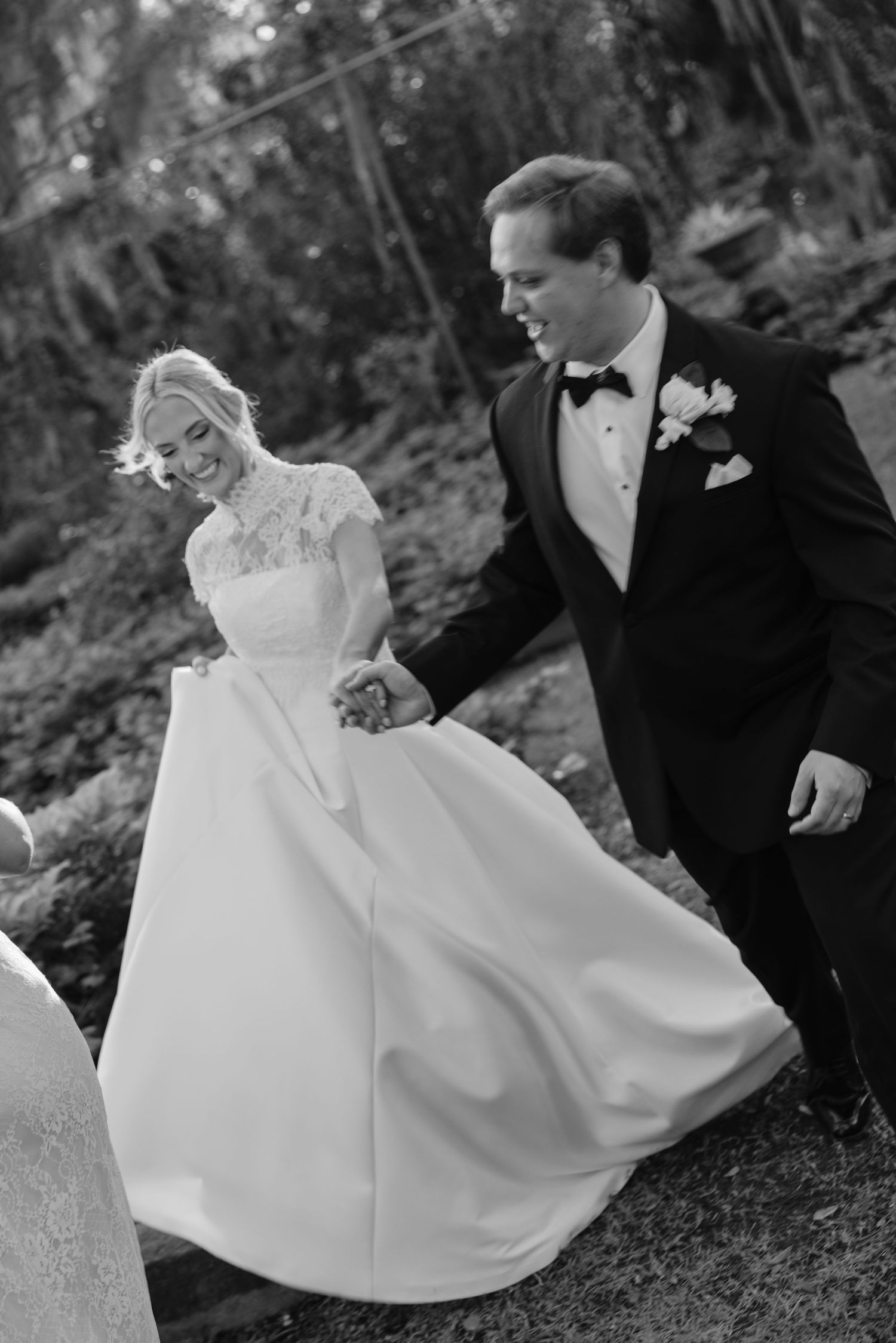 Black and white photo of a smiling bride and groom holding hands, walking outdoors in wedding attire.