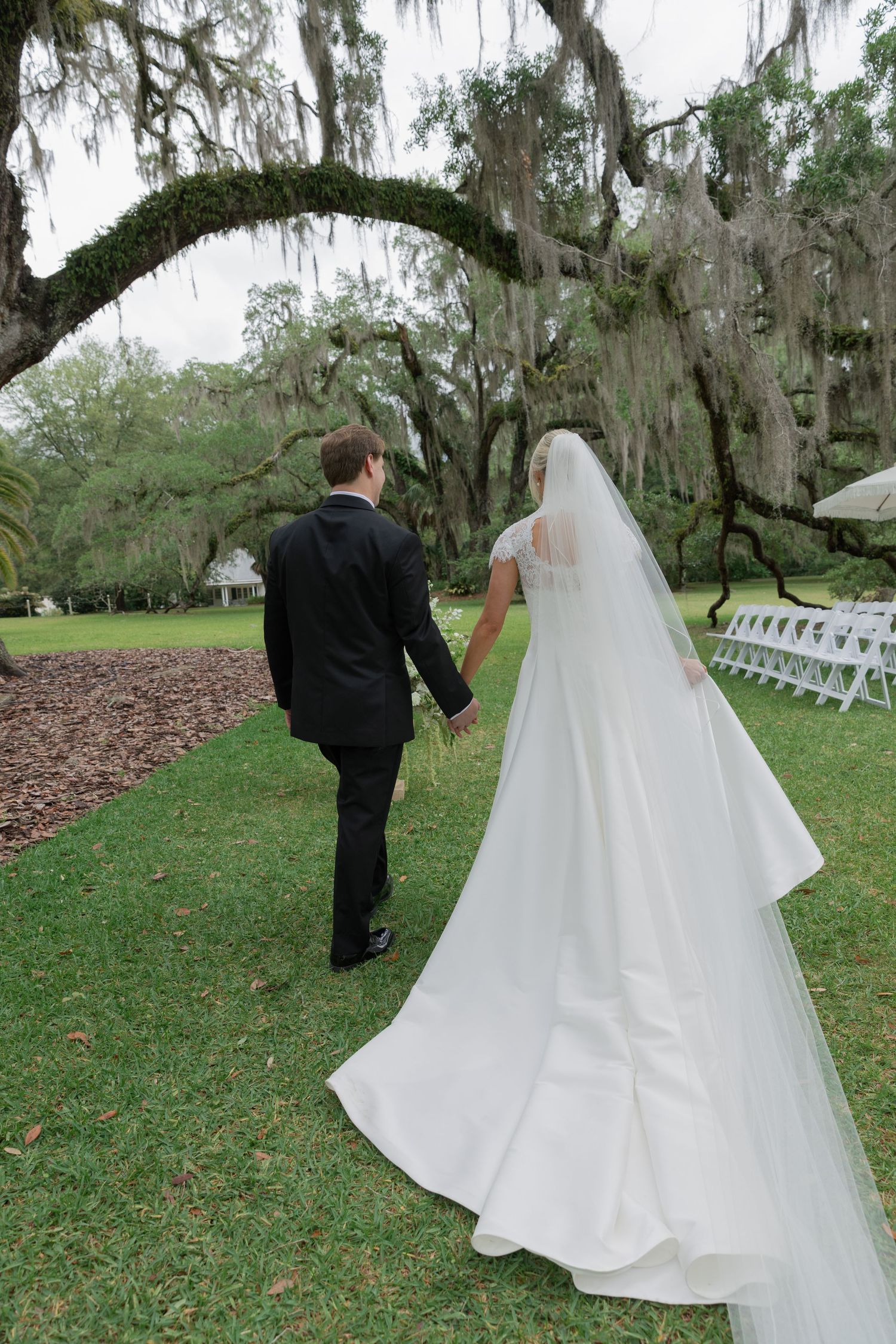 Bride and groom walk hand in hand across lush green lawn beneath moss-draped oak trees at outdoor wedding venue.