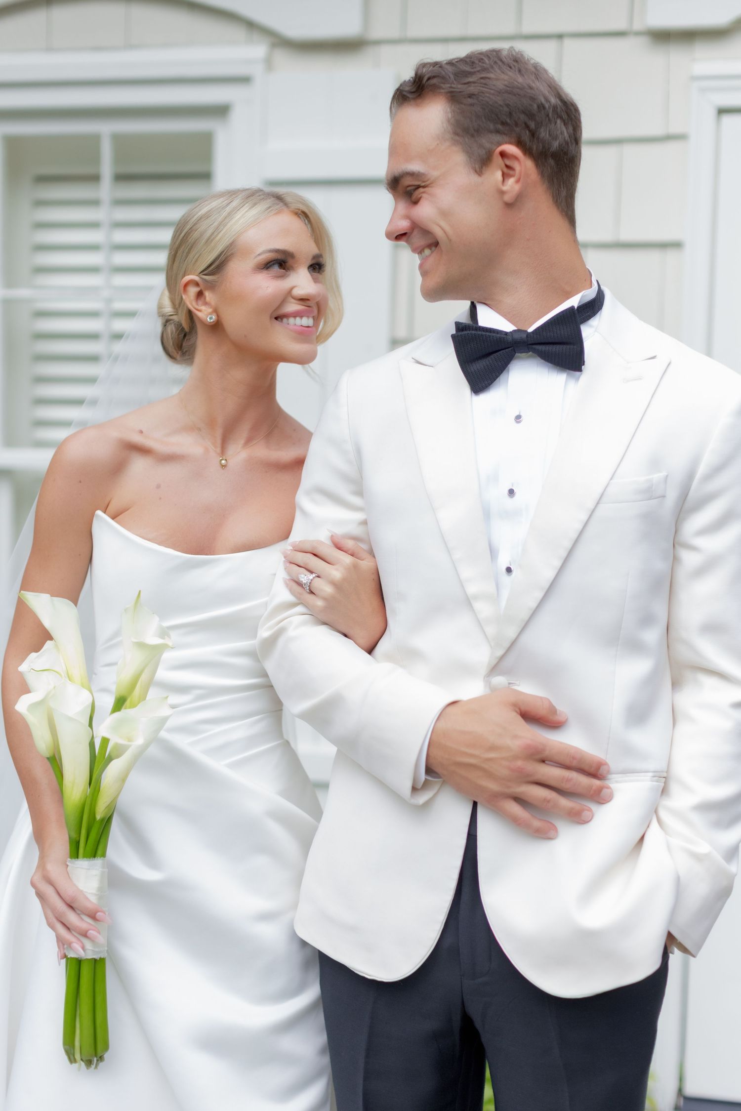 Smiling bride in a strapless satin gown holding white calla lilies links arms with a groom in a white dinner jacket and bow tie.
