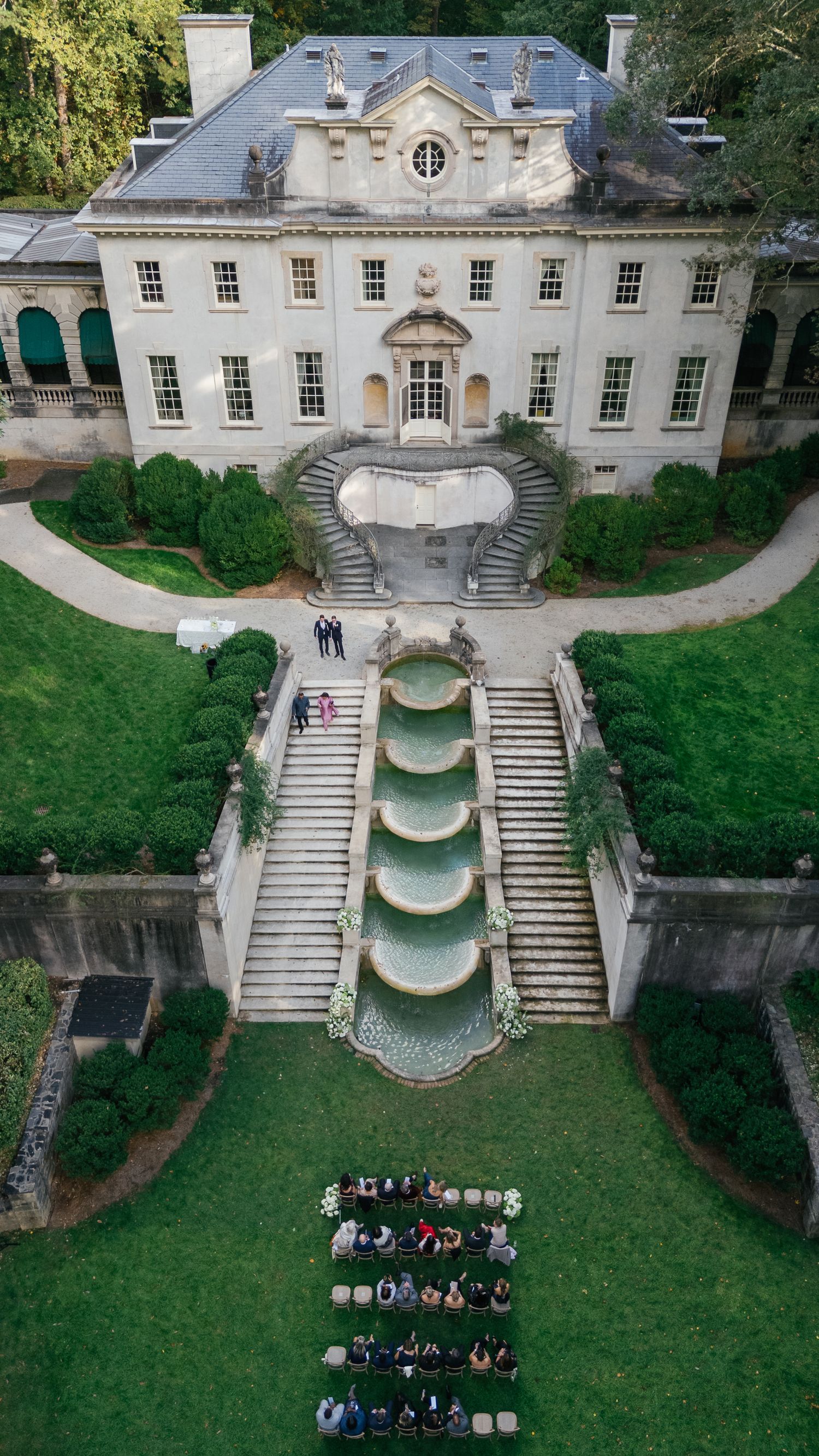 Aerial views of an elegant mansion with cascading fountains and curved stone steps where a wedding ceremony takes place.