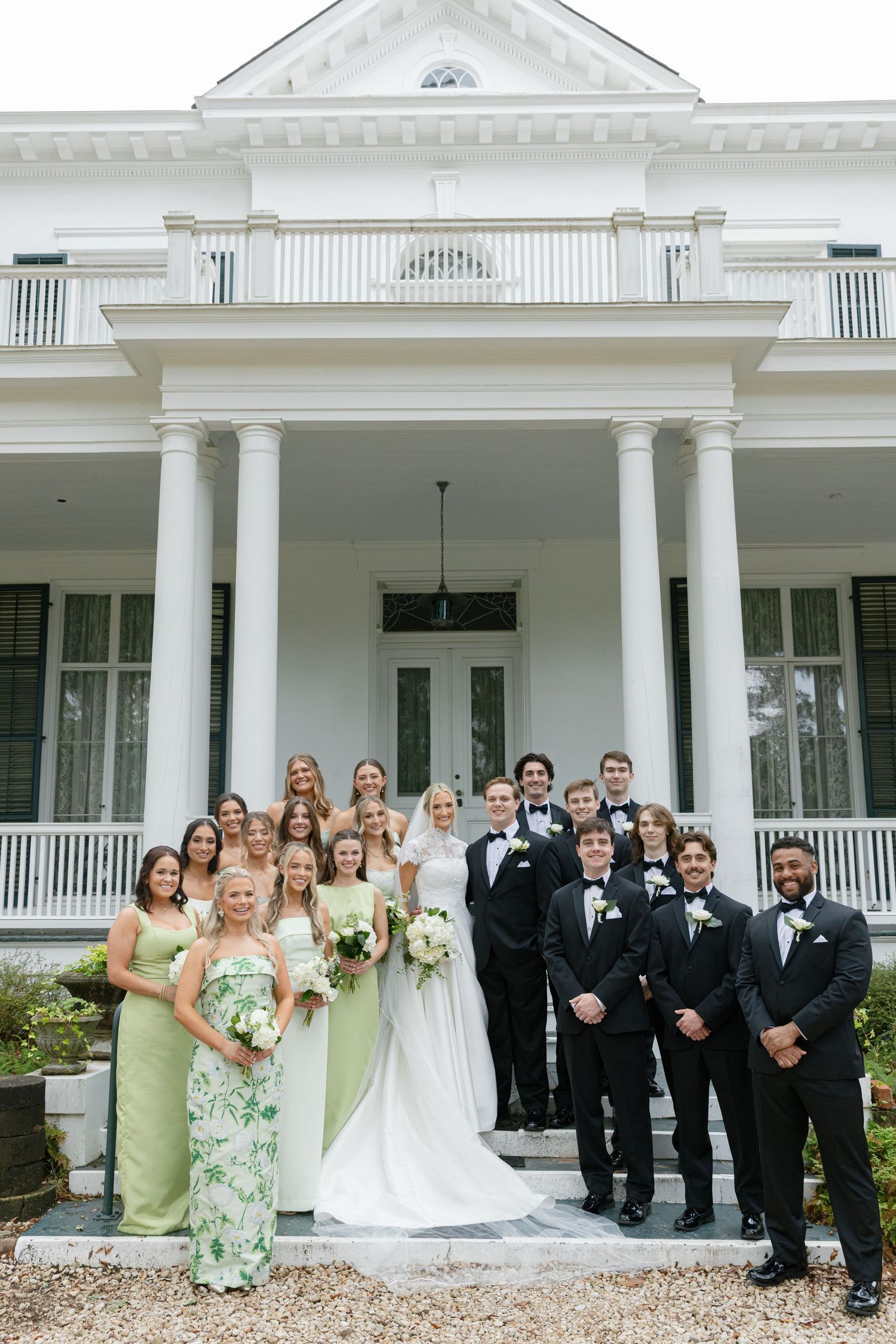 Wedding party poses on steps of grand white Southern plantation mansion with columns, bridesmaids in sage green gowns.