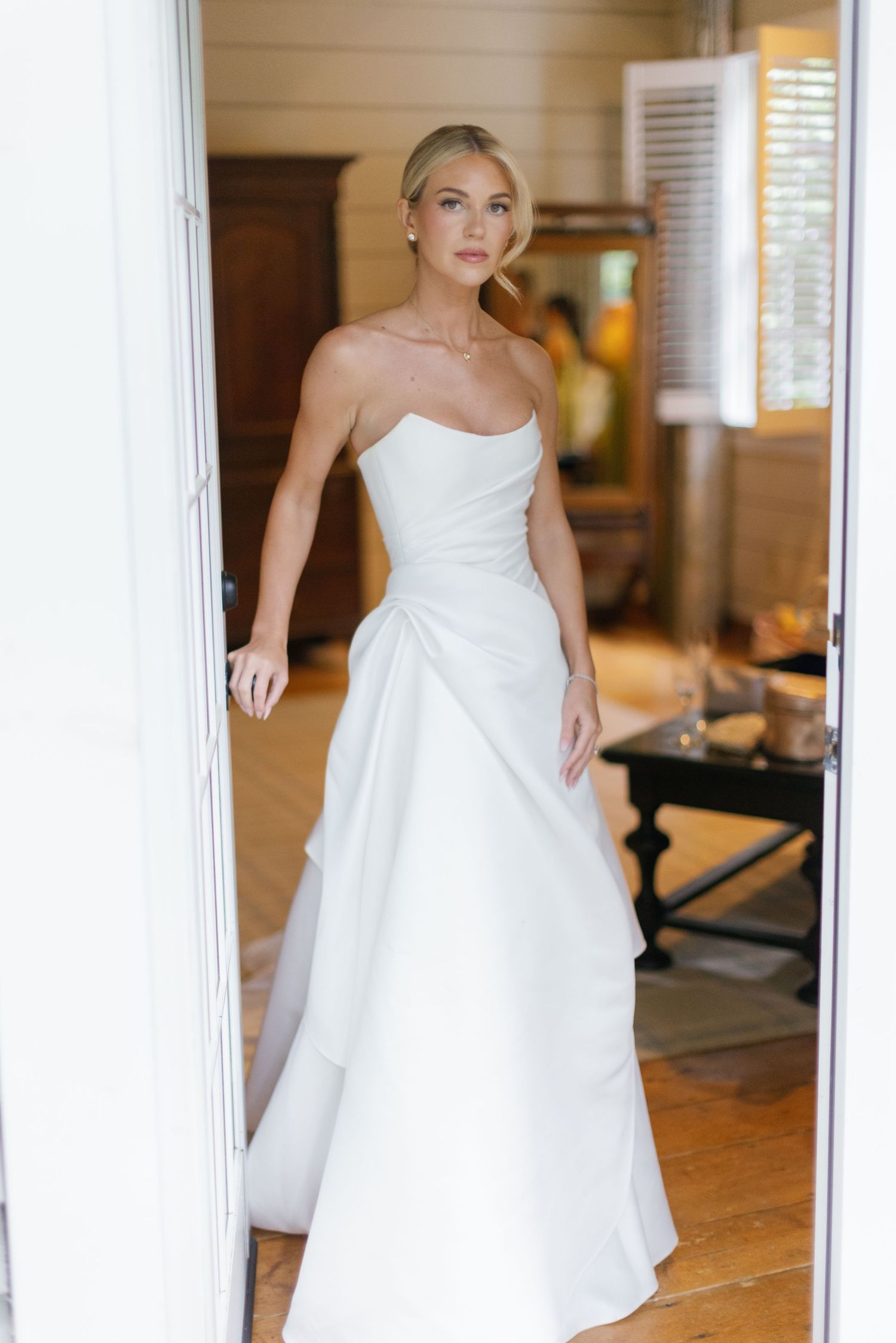 Bride in an elegant strapless white satin wedding gown with draped side detail stands in a sunlit open doorway of a rustic room.