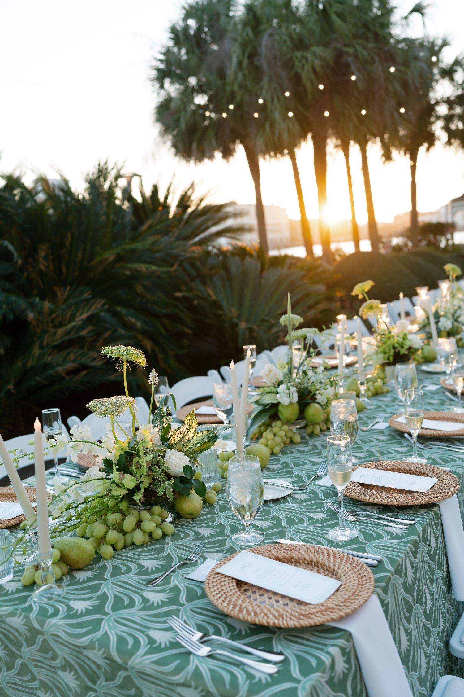 Elegant outdoor wedding reception table with green patterned linen, wicker chargers, floral centerpieces, and palm trees at sunset.