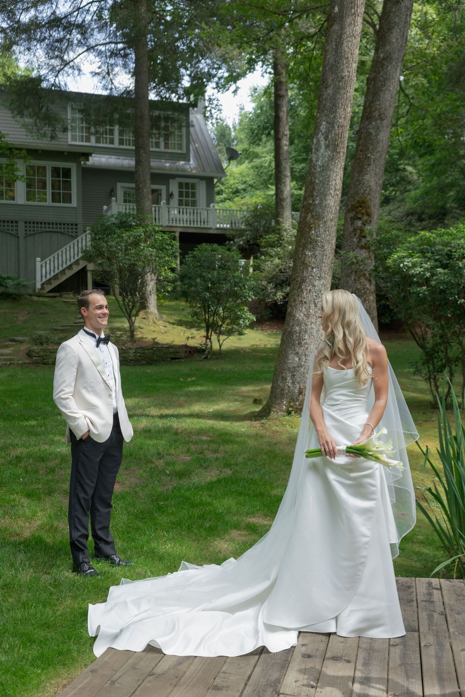 Bride in a flowing white gown and long veil holds a calla lily bouquet on a wooden deck as the groom in a white jacket watches nearby.