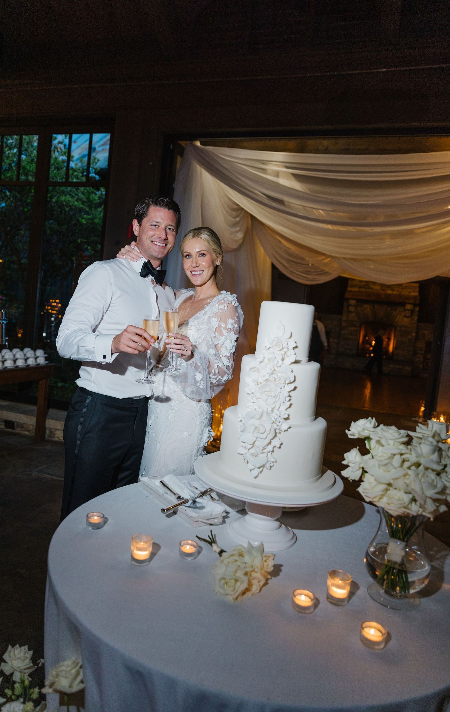 A wedding celebration with a couple toasting next to an elegant white tiered cake decorated with floral details.