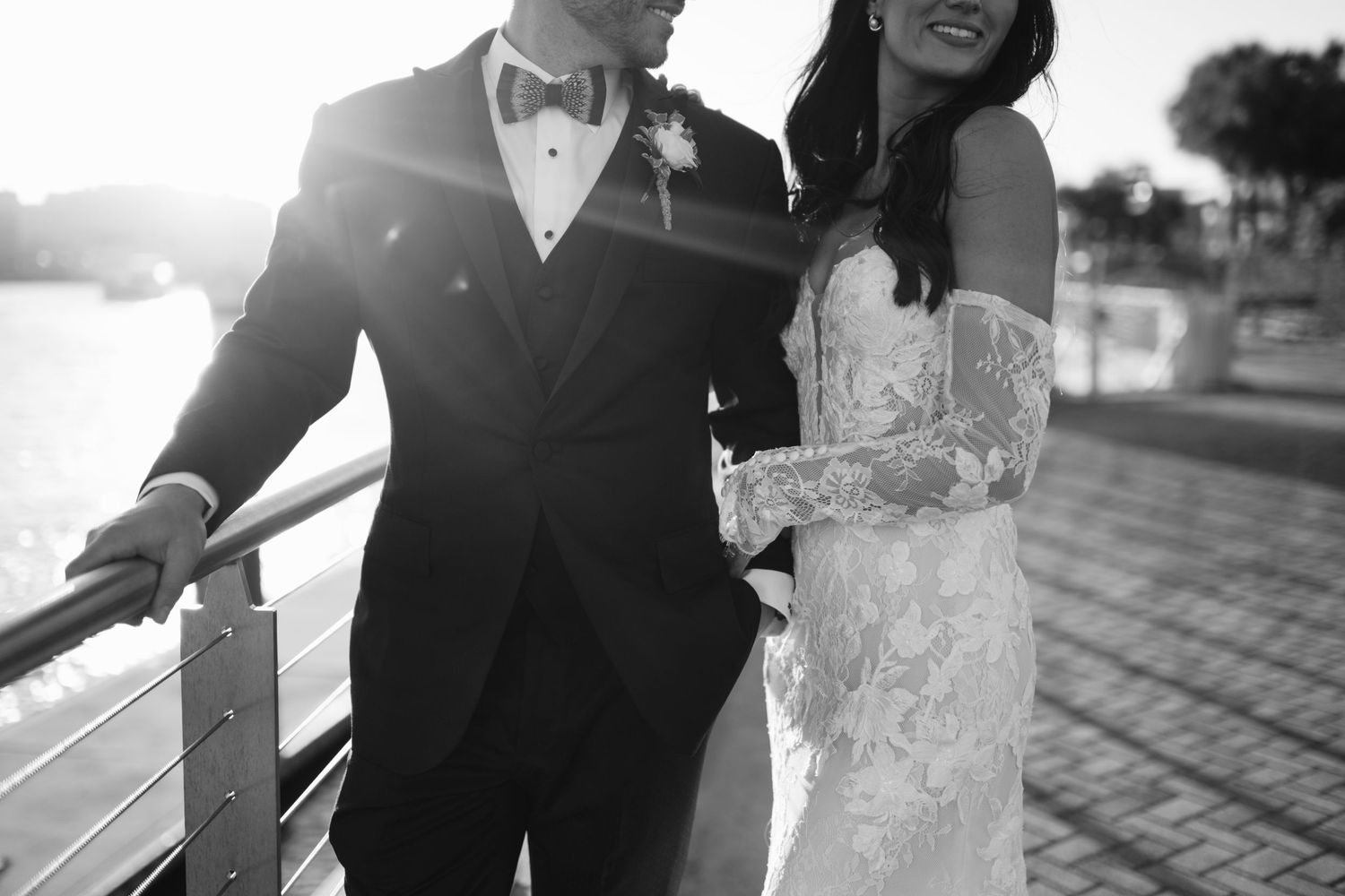 Black and white photo of a bride and groom standing together on a waterfront promenade at sunset.