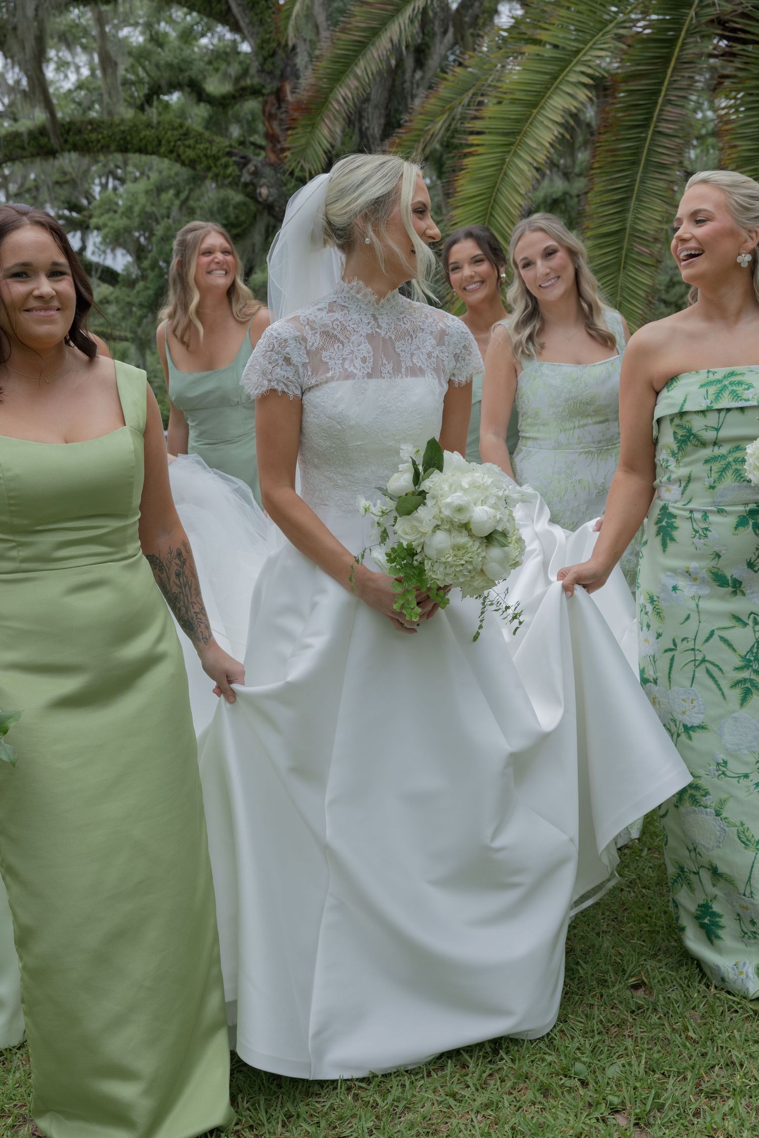 Bride in white lace gown holds bouquet surrounded by bridesmaids in green dresses outdoors.