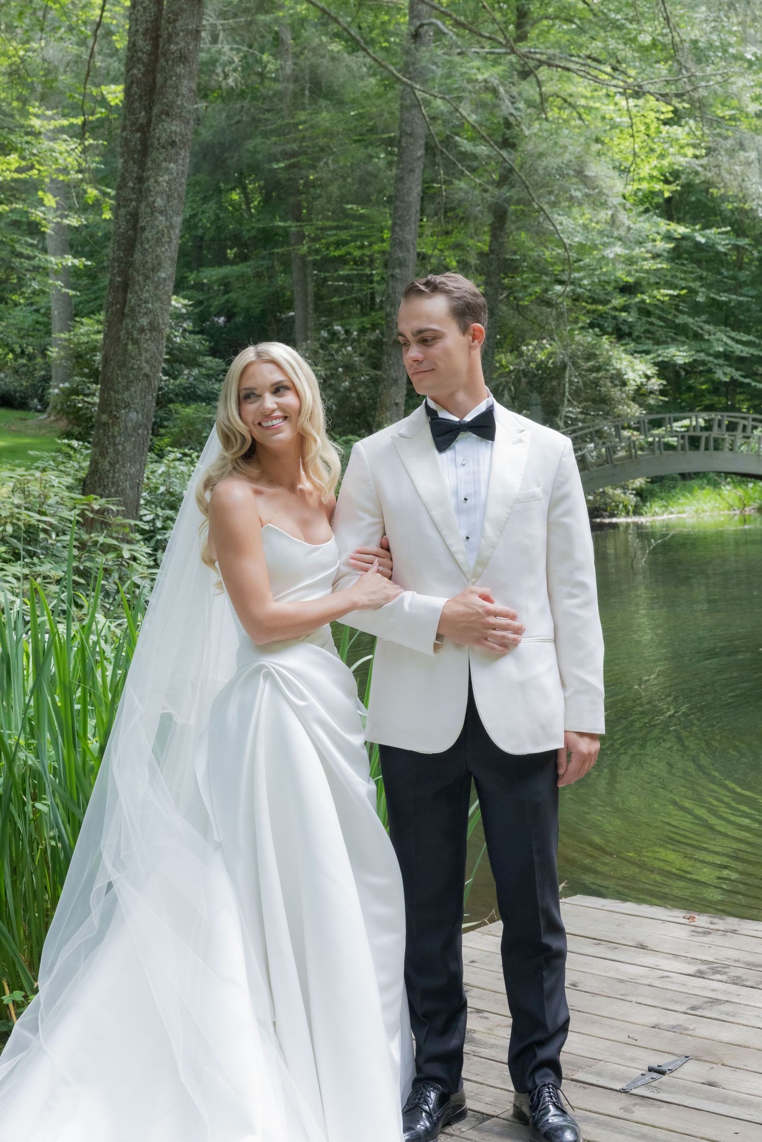 A bride in a white satin gown and veil stands beside a groom in a white dinner jacket on a wooden dock by a lake.