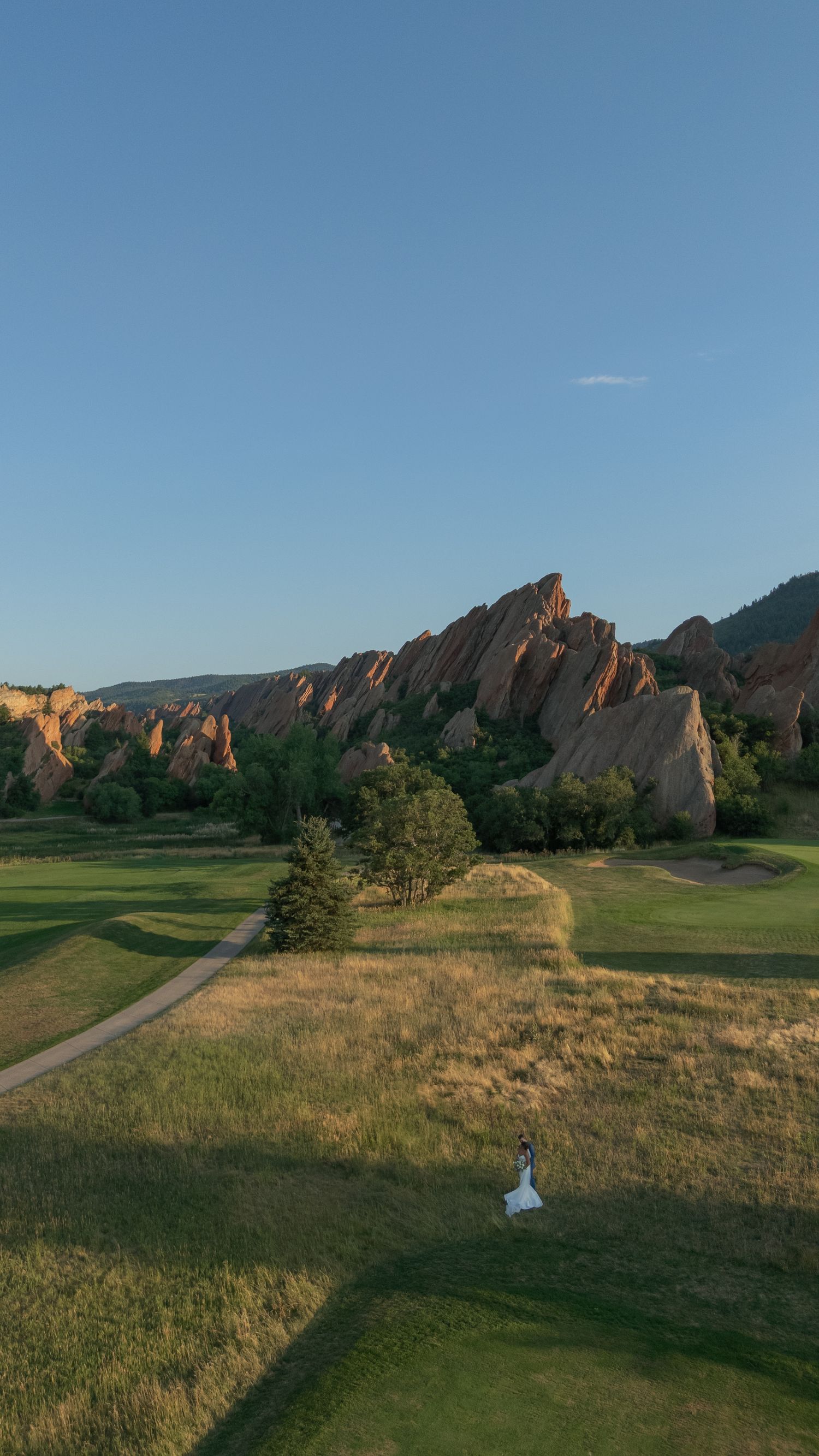 Aerial view of a bride in a white gown standing on a golf course fairway with red sandstone rock formations and a clear blue sky behind her.