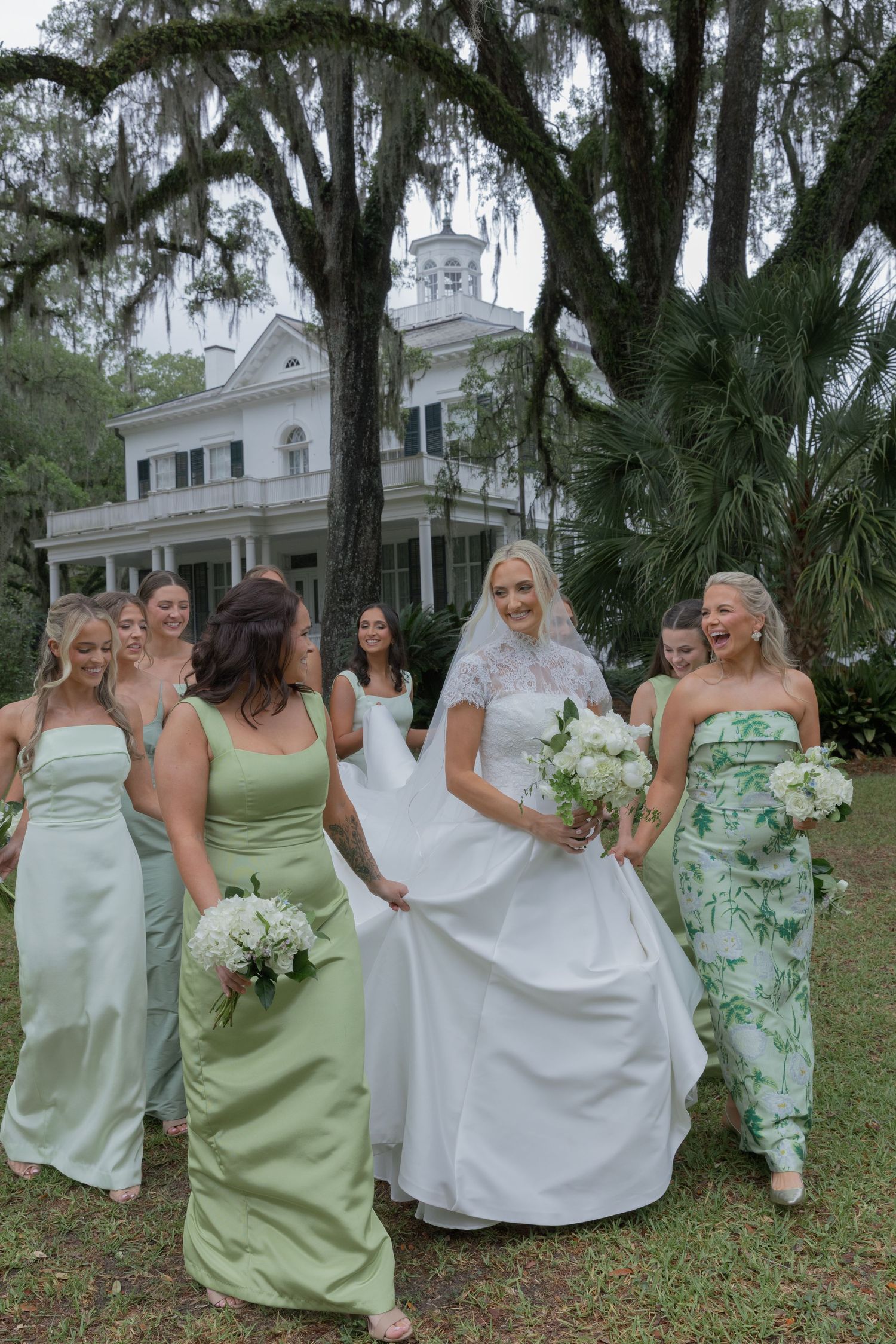 Smiling bride in white gown walks with bridesmaids in sage green dresses under Spanish moss trees at Southern estate.