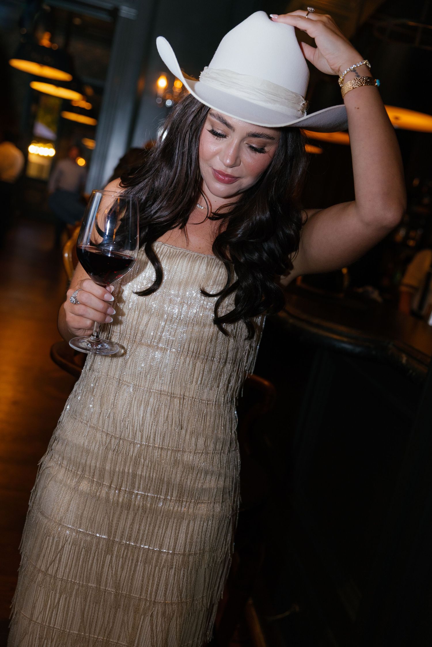 A woman in a sparkly dress and white cowboy hat holds a wine glass while dancing in a dimly lit venue.