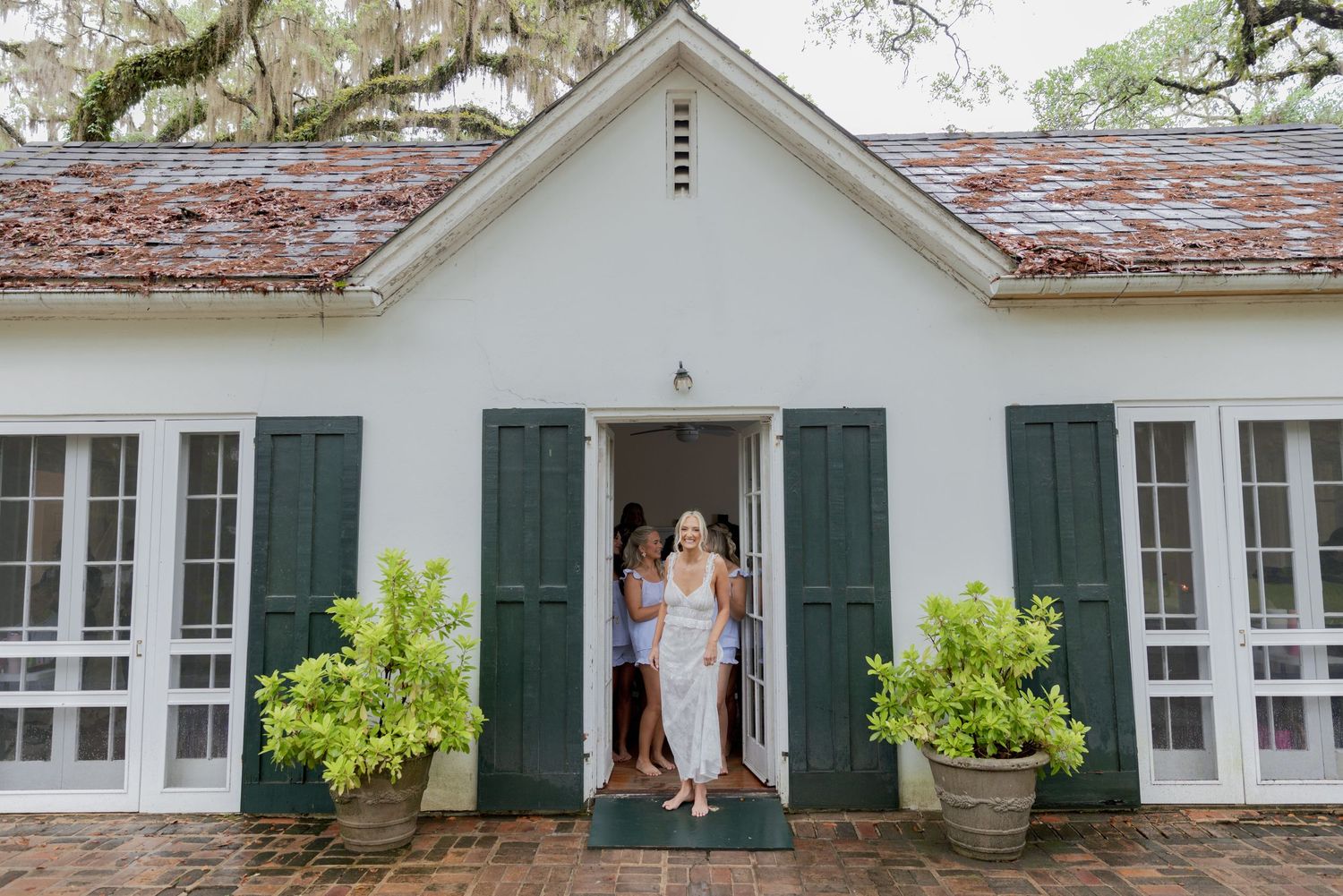 Two women stand in the doorway of a white cottage with green shutters and potted topiaries.