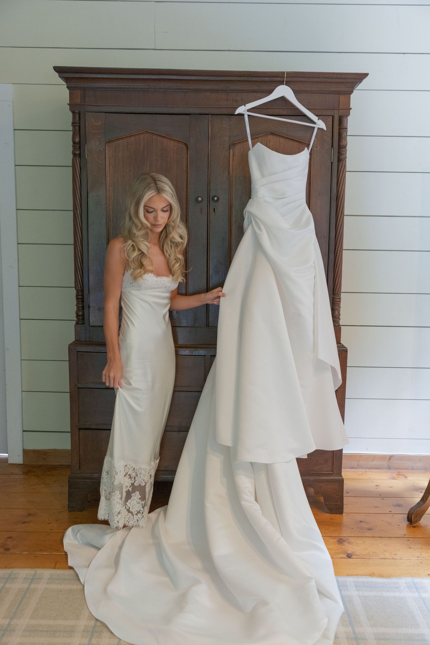 Bride in a fitted ivory gown touches her dress with a long train hanging on a hanger beside a rustic wooden armoire.