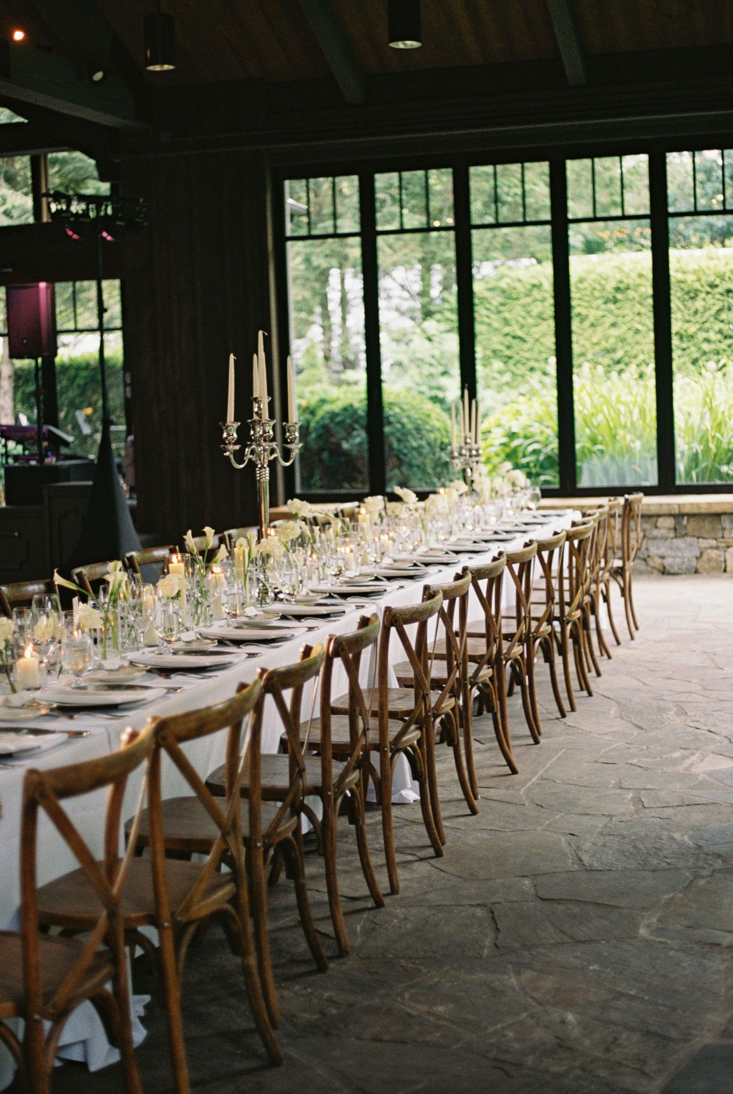 Long rustic wedding reception table with crossback chairs, candelabras and floral centerpieces in a stone-floored venue with garden views.