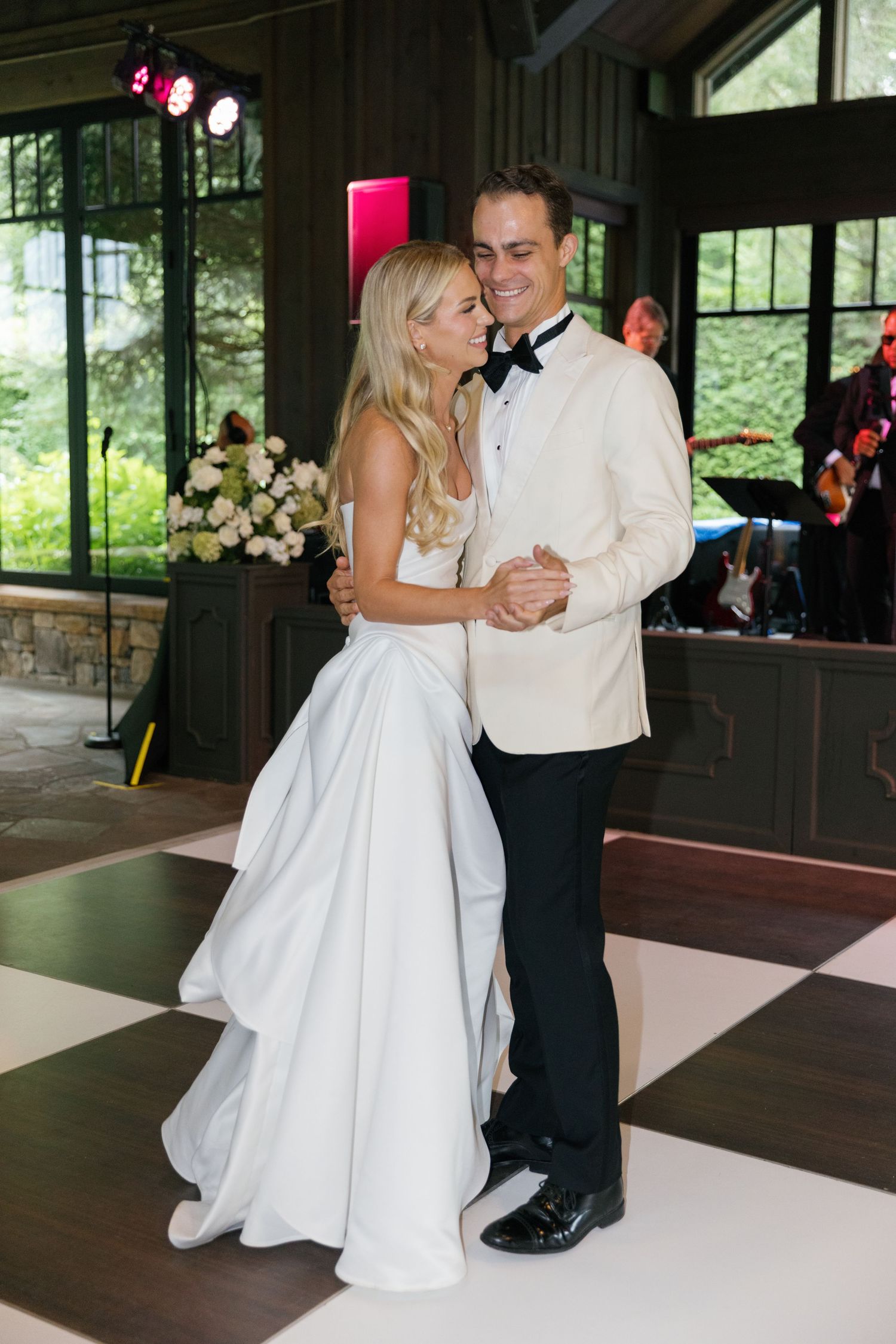 Smiling bride in a strapless white gown and groom in a cream dinner jacket and bow tie share a first dance on a checkered floor.